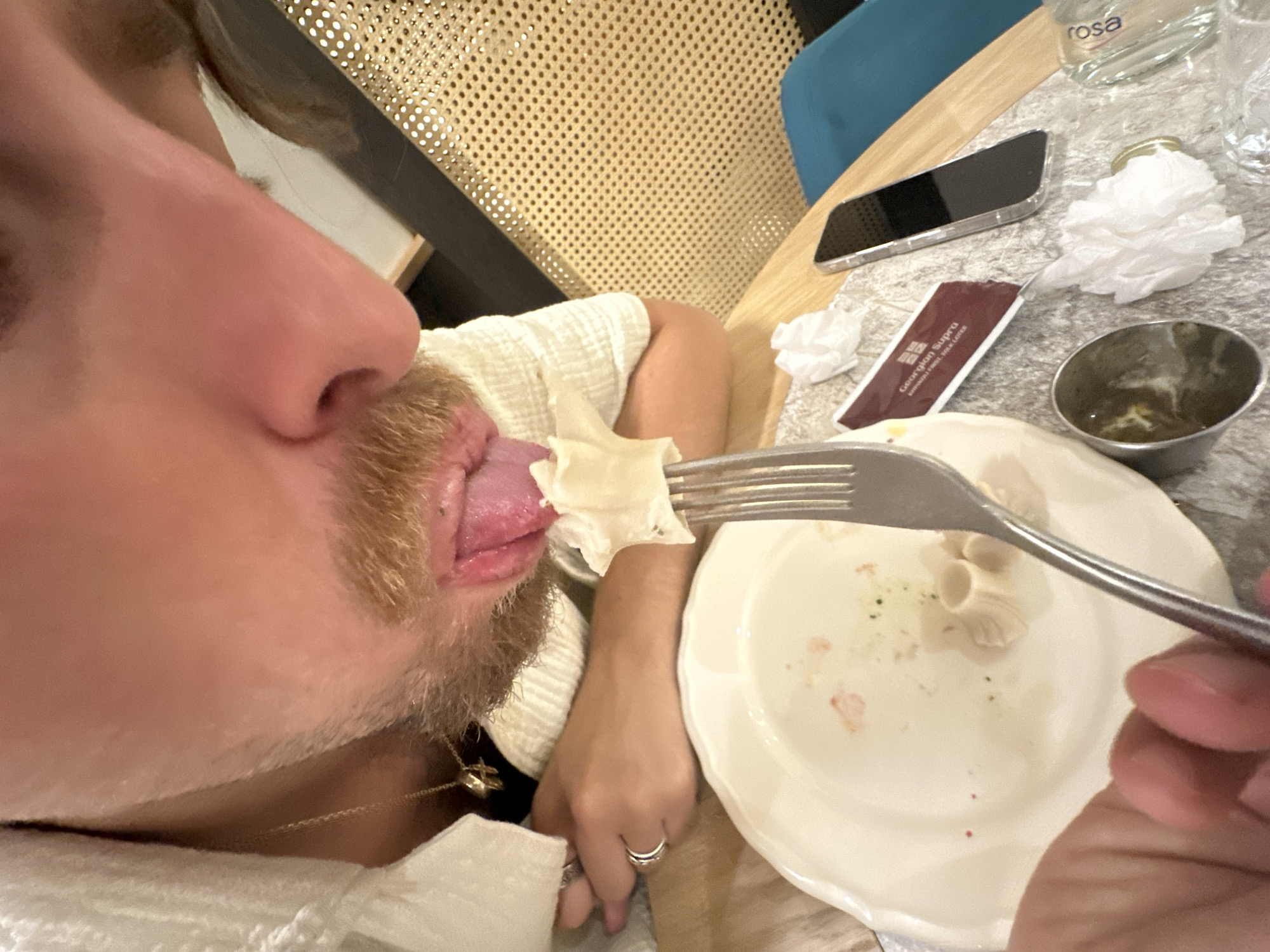 The image shows a close-up of a person's face and mouth, with their tongue extended, about to lick food off a fork. The person has light brown facial hair and is wearing a light-colored shirt and a gold necklace. A partially eaten meal is on a white plate in front of them. The scene appears to be indoors, possibly a restaurant, with a table made of wood and a woven cane chair in the background. A smartphone, a small metal container, and a perfume bottle are visible on the table. The location is specified as Belgrade, Serbia.