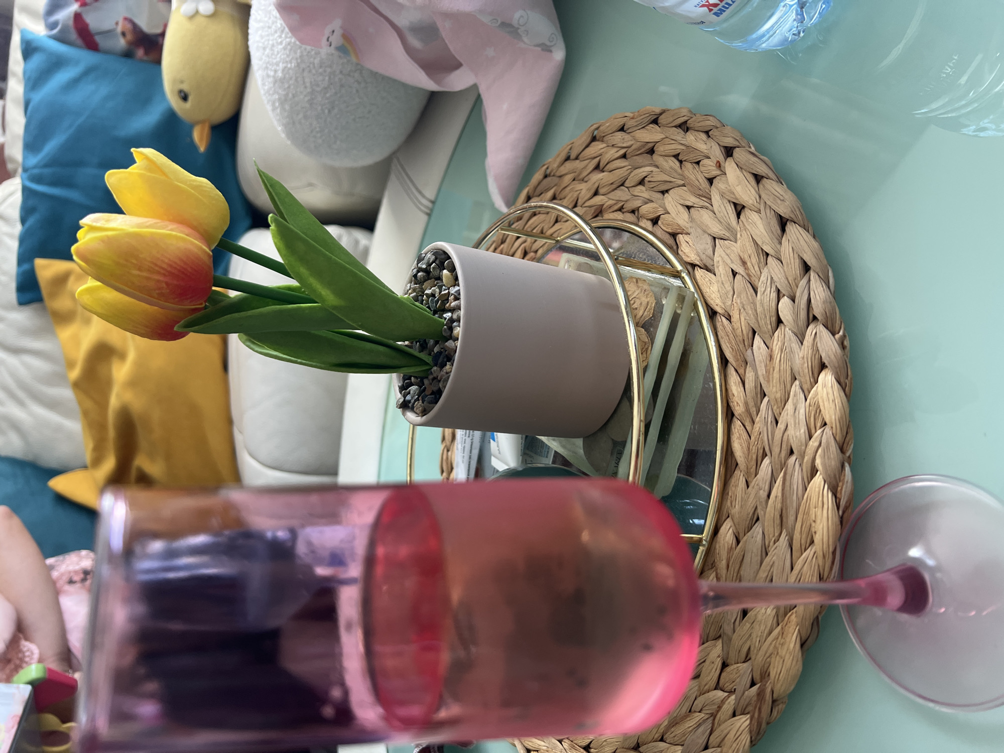 The image depicts a still life arrangement on a light green surface. A pink wine glass dominates the foreground, its form obscuring some of the other elements. Behind the glass, there's a small potted plant with yellow and orange tulip blossoms and green leaves. The plant sits in a light beige pot filled with dark pebbles. The pot rests on a circular straw mat, which in turn sits on a gold-framed mirror stand. In the background, there is a cluttered space with pillows in various colors (blue, yellow, white) and a stuffed animal. The image appears to have been taken indoors, and there is no discernible information to confirm this image was taken in Plovdiv, Bulgaria. The presence of the pink wine glass and the other objects suggests an informal, domestic setting. No people are fully visible, though a small part of a person is seen on the left.