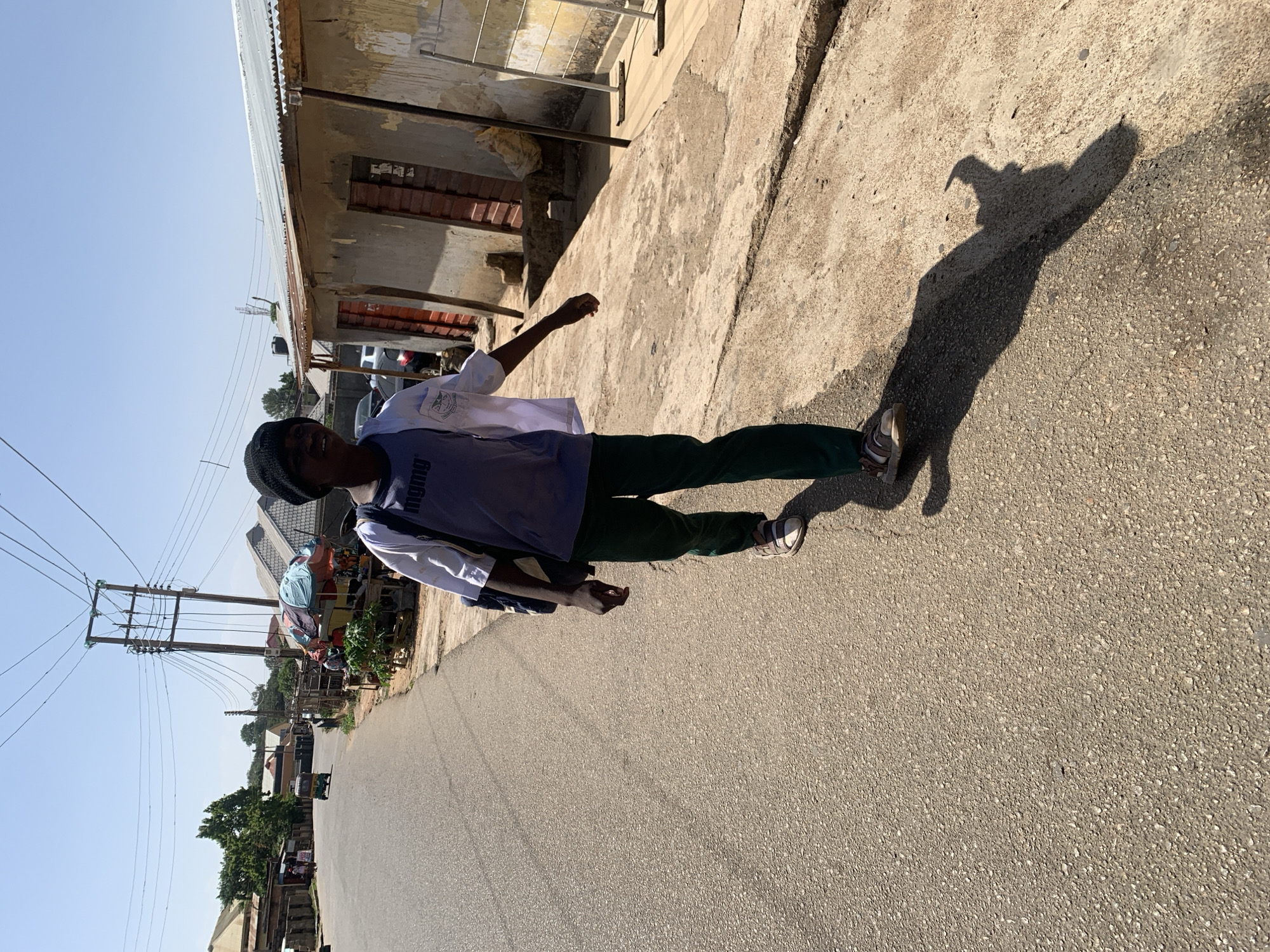 The photo shows a young person standing on a paved road in Jos, Nigeria. The person is wearing a white shirt with a blue vest, green pants, and sandals. They are casting a shadow on the road. In the background, there are buildings, power lines, and trees. A motorcycle is seen in the distance.