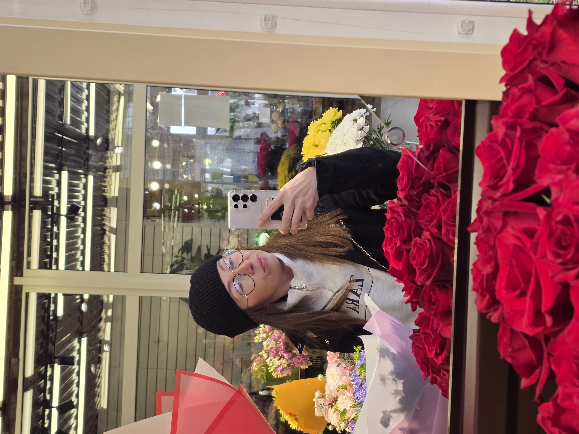 A person with long brown hair, wearing a black beanie and round glasses, takes a selfie in a mirror. The person is holding a white smartphone. The reflection shows a variety of flowers, including red roses, yellow and white daisies, and bouquets with pink and purple flowers. The background visible in the reflection suggests an indoor setting, likely a flower shop. The image is rotated 90 degrees clockwise. No information is available about the city of Chaikovsky, Russia.