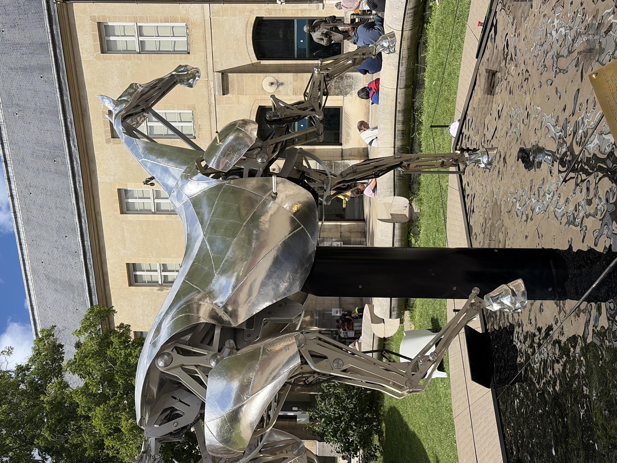 A large, metallic sculpture of a horse stands in what appears to be an outdoor courtyard or plaza. The sculpture is highly reflective, mirroring the surrounding environment. It is intricately detailed, showcasing mechanical components within its structure. The horse sculpture is positioned over a dark, reflective surface, possibly a fountain or pool, with ripples visible on its surface.
In the background, a light-colored building with a grey roof and multiple windows is visible. A large tree with green foliage is to the left of the sculpture. Several people are present in the background, some sitting on a curved stone bench or wall, while others appear to be standing. One person is visible in the distance, standing near a statue. The ground around the sculpture is covered in grass and a light-colored, textured walkway. The sky is blue with some clouds.
The scene is likely located in Paris, France, given the architectural style of the building and the presence of public art installations. This specific artwork is likely a modern kinetic sculpture, given its complex mechanical appearance. The sculpture is positioned in a public space, suggesting it is an art installation for viewing and appreciation.