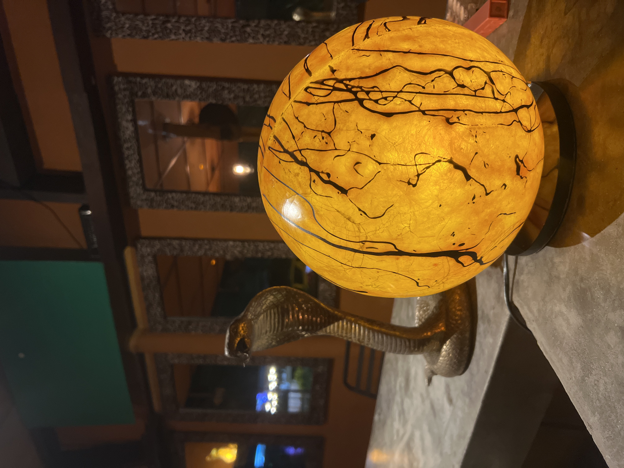 A lit lamp with a globe shade rests on a stand shaped like a cobra. The globe is an orange-yellow color with black patterns. The cobra stand is silver-colored and appears to be made of metal. The lamp is positioned on a gray surface that seems like a ledge or counter. In the background are wall mirrors with reflections of the surrounding environment, including what looks like interior lighting and a glimpse of a green wall. Based on the information provided, the location is Baan Lamai, Thailand. The exact setting is indoors, likely a bar or restaurant based on the background reflections.
