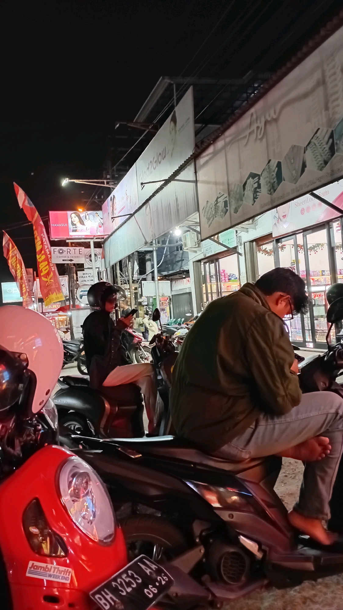 In Jambi, Indonesia, at night, several individuals are gathered on motorcycles in front of shops lining a street. One person, viewed from the back, wears a green jacket and gray pants, with bare feet resting on a dark motorcycle. Another person in the foreground, wearing a black jacket and helmet, is looking at their phone while seated on a dark motorcycle. A third person, also in a helmet, is sitting on the same motorcycle as the person with the phone. A red motorcycle is parked in the foreground on the left, with a white helmet on top. Billboards and signs are visible above the shops, including one advertising "Glow2Glow" and another with "FORTE". The street appears to be illuminated by artificial lights.