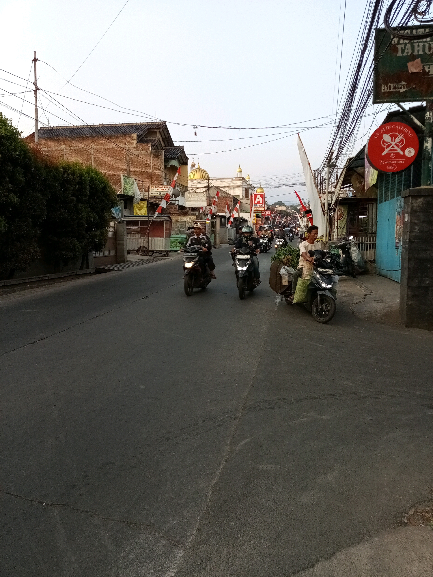 A street scene in Sariwangi, Indonesia, features a paved road with several motorcycles in motion. On the right side of the road, a blue building is visible, next to a brick building with a dark tiled roof. A traffic pole with multiple wires is positioned on the left side of the road, near a large tree. Several shops line the street, with a red circular sign advertising "CV. ALDI CATERING" prominently displayed on the right. Red and white flags are mounted on poles along the roadside. One motorcycle, driven by a man in a white shirt and a cap, carries a large sack of green produce. Another motorcycle, ridden by a person wearing a helmet, is also on the road. The sky above is clear and bright, suggesting it is daytime.