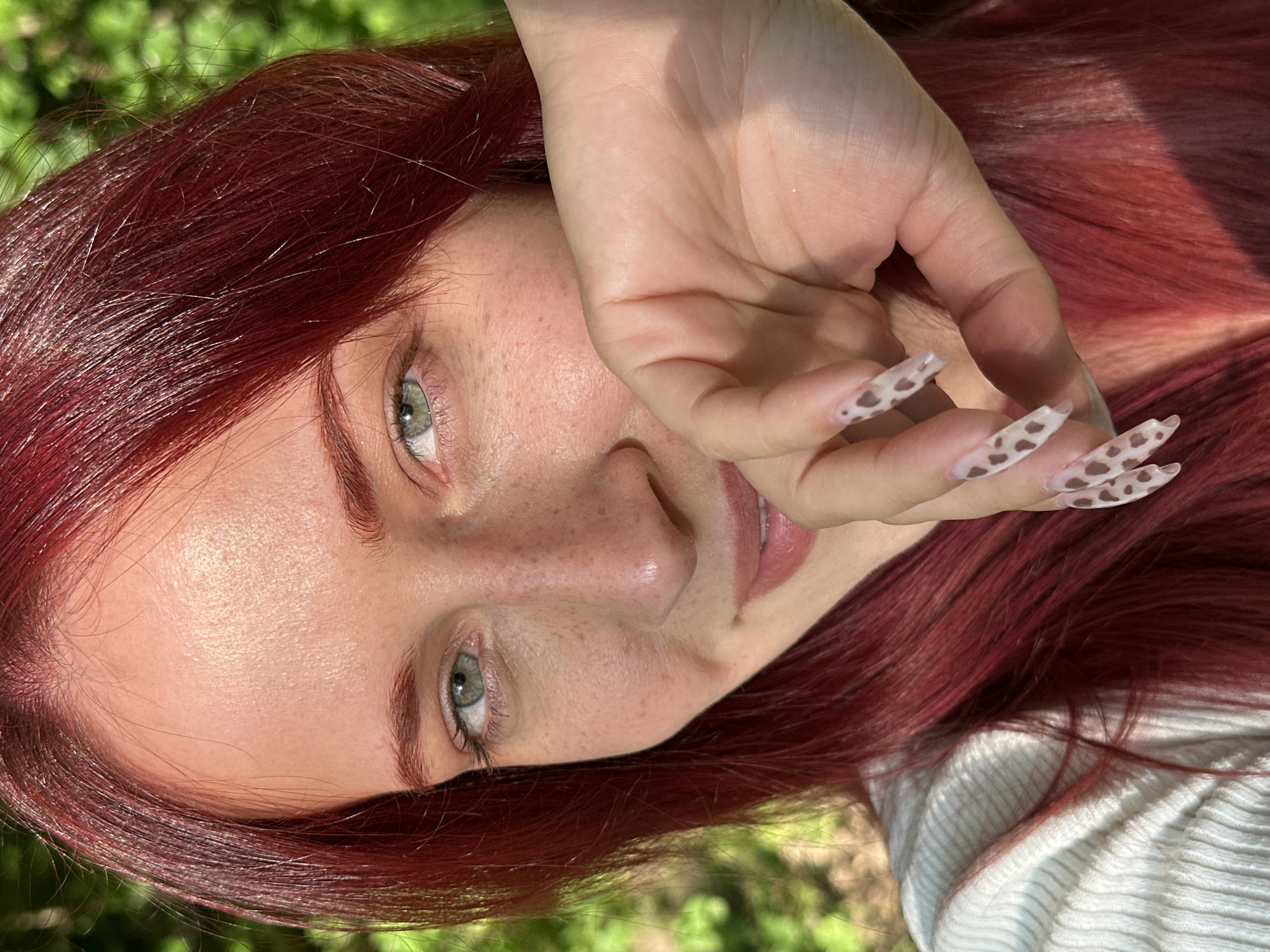 A close-up, brightly lit portrait features an adult female with long, deep auburn hair and light green eyes, looking directly at the viewer. Her fair skin displays numerous freckles across the nose, cheeks, and under-eye area. A subtle smile is visible on the upper part of her face, while her lower mouth and chin are partially obscured by her raised left hand. The fingernails on this hand are long, shaped into a tapered square or coffin style, with a translucent base and irregular dark brown spots, resembling an animal print. She is wearing a light-colored, ribbed knit top. The background is a uniformly soft-focus expanse of vibrant green foliage, indicating an outdoor natural setting. Strong, direct sunlight illuminates the scene, casting highlights on her hair, forehead, nose, and the back of her hand, and creating subtle shadows. The image provides no discernable details to identify the location as Moscow, Russia.