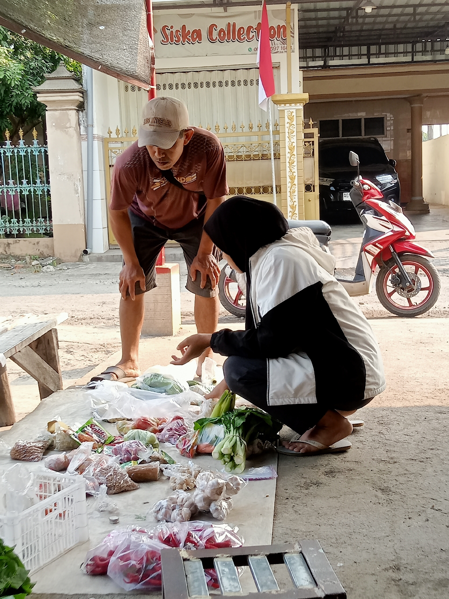 This image depicts a street-level vendor selling produce in Simo, Indonesia. Two individuals are present: a man wearing a maroon t-shirt and a baseball cap, and a person wearing a black and white hooded jacket and a headscarf. The man is bending over, looking down at the goods displayed on a table. The person in the jacket is crouched, appearing to be selecting or interacting with the produce.
The scene is set outdoors, with a building visible in the background bearing a sign that reads "Siska Collection." A red and white Indonesian flag is attached to a pole next to the building. Various types of produce are spread out on a tarp or cloth-covered surface, including leafy vegetables, chilies, garlic, and packaged goods. A white plastic crate is also visible. A black car and a red and white motorcycle are parked behind the vendor's display, suggesting this is a roadside market. The ground is paved with dirt or gravel. The lighting suggests it is daytime, with sunlight casting shadows.