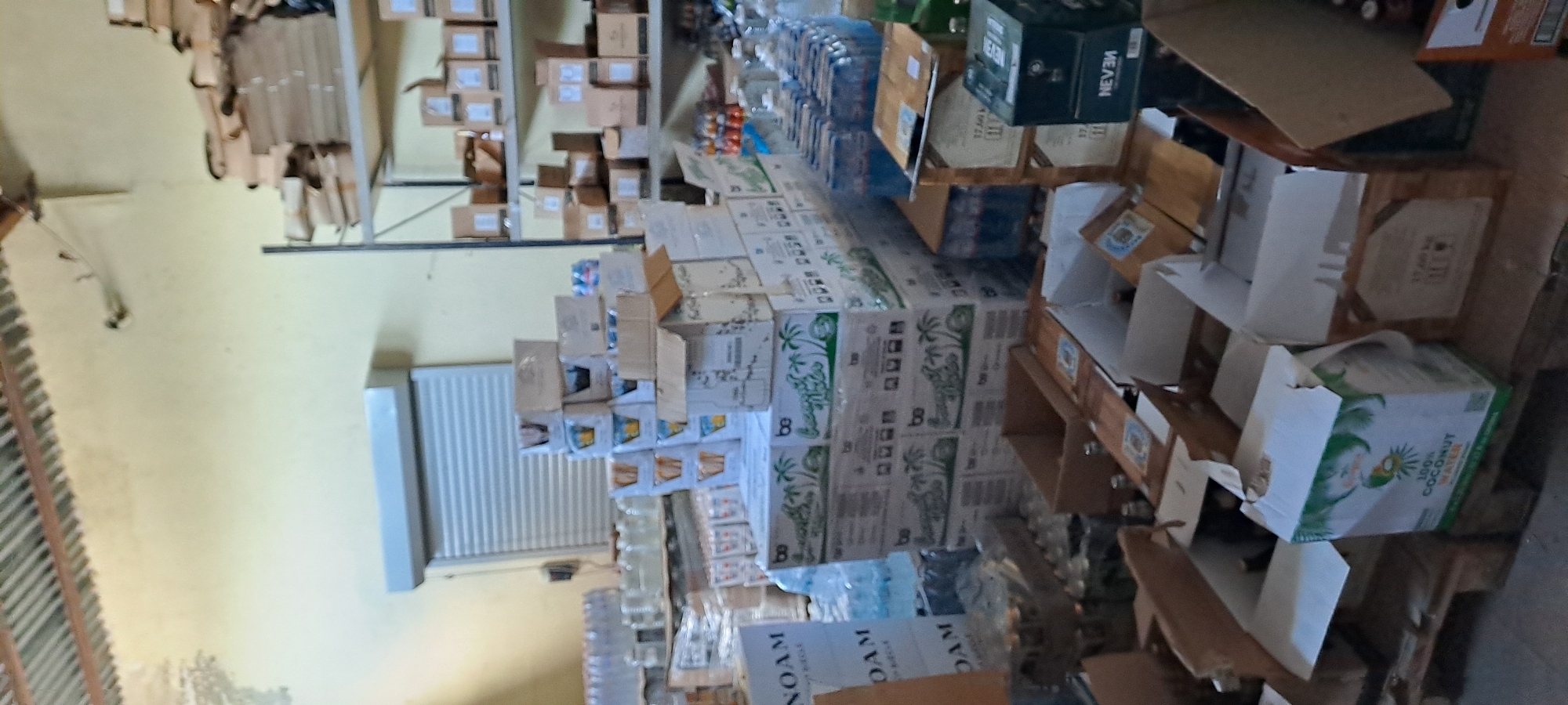 The image depicts a warehouse interior with stacks of boxes and bottled water. In the foreground, several cardboard boxes are arranged on wooden pallets. One box prominently displays "100% COCONUT WATER" with an image of a coconut. Other boxes are partially visible, some open and containing bottles. To the left, stacks of water bottles are wrapped in plastic. In the mid-ground, more boxes, some with branding that includes "be," are stacked high. In the background, shelves are laden with more boxes. A grey roll-up door is visible on the left wall, and the ceiling has corrugated metal sheeting. The overall environment appears to be a storage or distribution facility. No people are present in the image. The location is identified as Velika, Bulgaria.