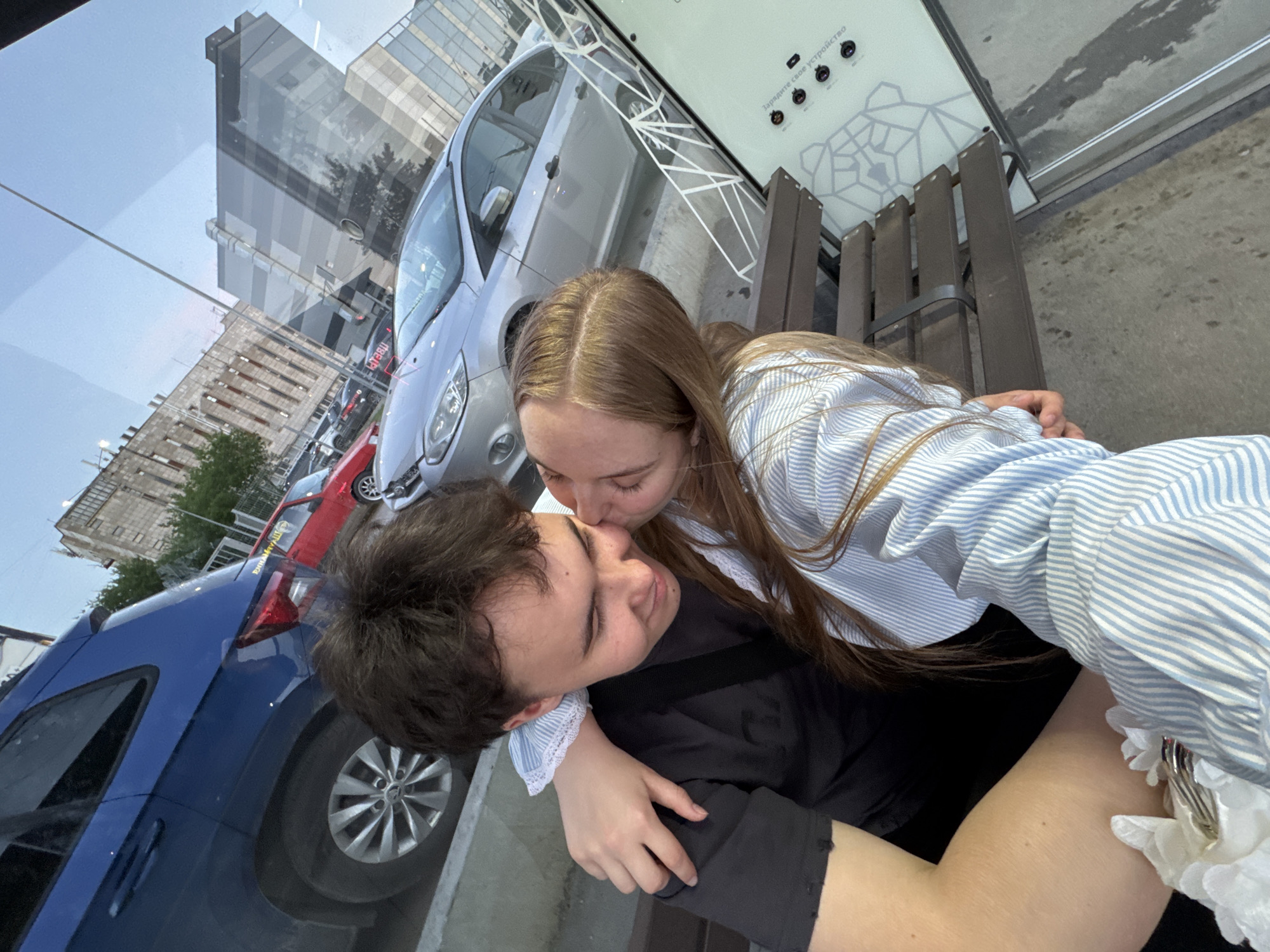 A couple embraces in Perm, Russia. The female, fair-skinned with long blonde hair, kisses the male, who has dark hair. She is wearing a light blue and white striped shirt; he is wearing a dark shirt. They appear to be seated on a bench in an urban environment. Reflected in a glass surface nearby are buildings, parked cars (blue, silver, and red), and possibly a map display with electronic devices (possibly cameras).