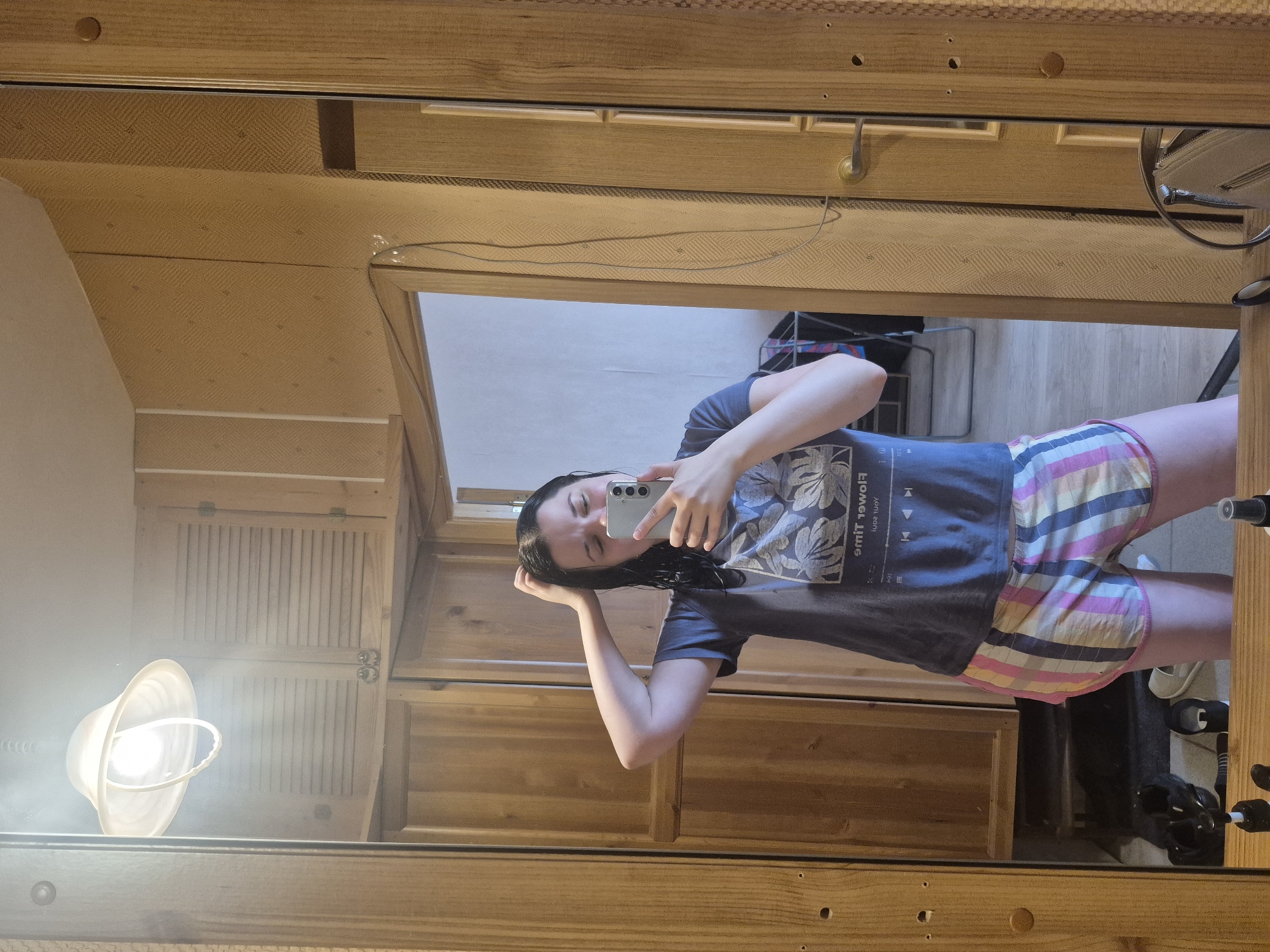 A young woman with dark hair, wet from showering, stands in front of a mirror and takes a selfie. She is wearing a dark grey t-shirt with a floral design and multi-colored plaid shorts. Her left hand is raised and touching her head, while her right hand holds a silver smartphone. The mirror reflects a room with wood-paneled walls and cabinets. A ceiling lamp with a white shade is visible in the reflection. In the background, through an open doorway, a clothes rack with laundry can be seen. The floor is tiled, and a pair of black sneakers and white sandals are on the floor. The scene appears to be in a bathroom or dressing area within a residential setting. The location is not identifiable as Saint Petersburg, Russia.