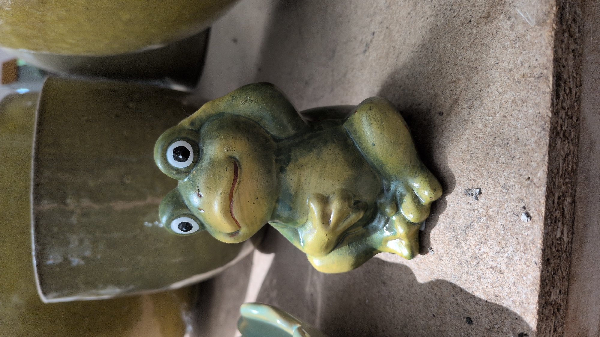 This is a close-up, eye-level, outdoor shot of a green ceramic frog statue sitting on a rough, light-brown surface. The frog is in a seated position, with its hands clasped in front of it and its head tilted slightly to its right. It has large, round, white eyes with black pupils, and a small, reddish mouth. Behind the frog, out of focus, are large, dull green ceramic pots. The surface the frog is sitting on appears to be a wooden plank or shelf, with some small debris scattered on it. The lighting suggests a sunny day, casting a shadow of the frog onto the surface. There is no indication of the specific location within Sömmerda, Germany, but the scene suggests an outdoor garden or pottery display.