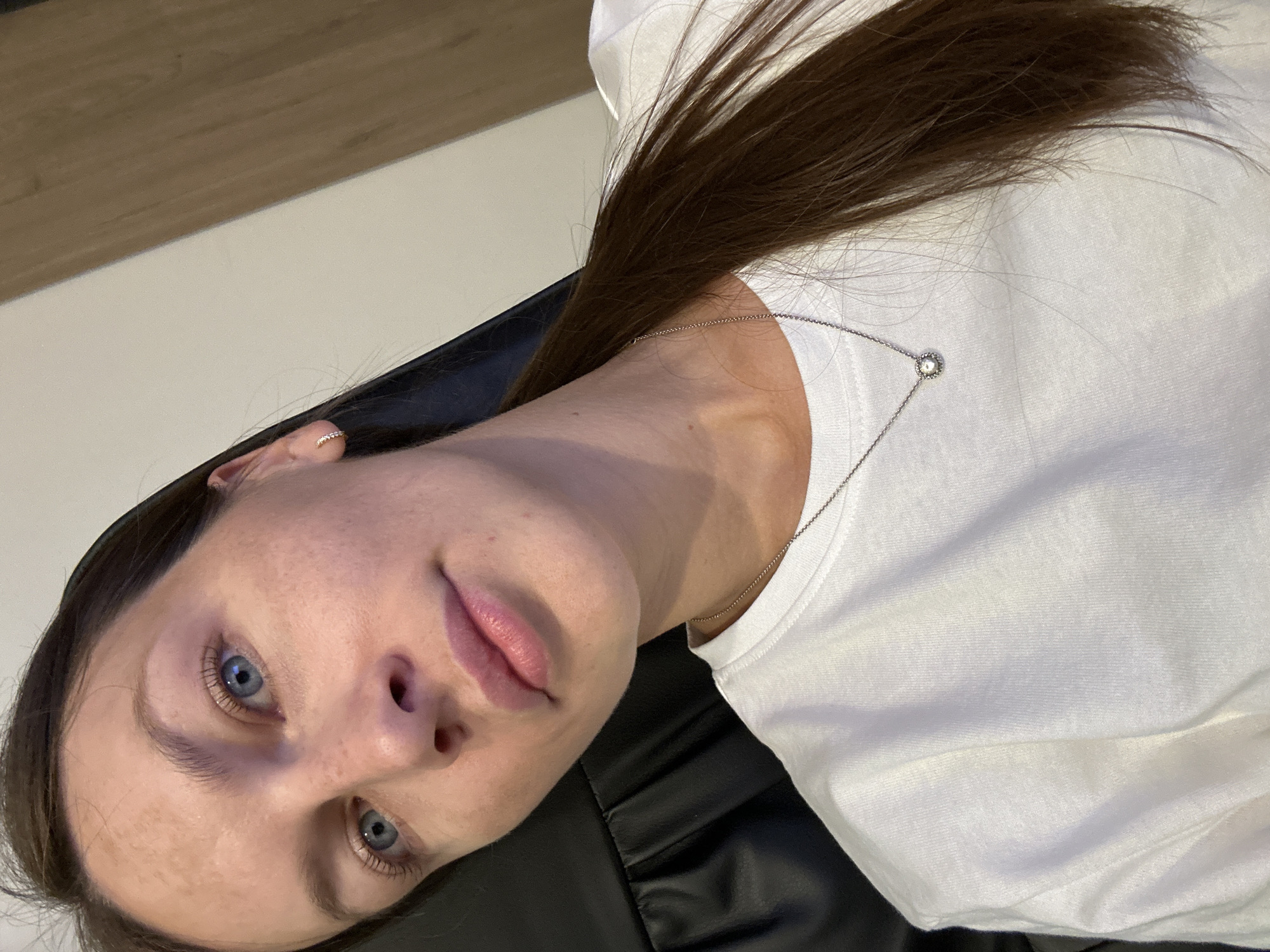 A woman with brown hair and blue eyes is shown from the chest up, wearing a white t-shirt and a silver necklace with a diamond pendant. She is seated in a black chair, with a wooden surface visible in the background. The lighting is natural, and the image appears to be taken indoors. No specific location in Boreti, Montenegro is identifiable from the image.