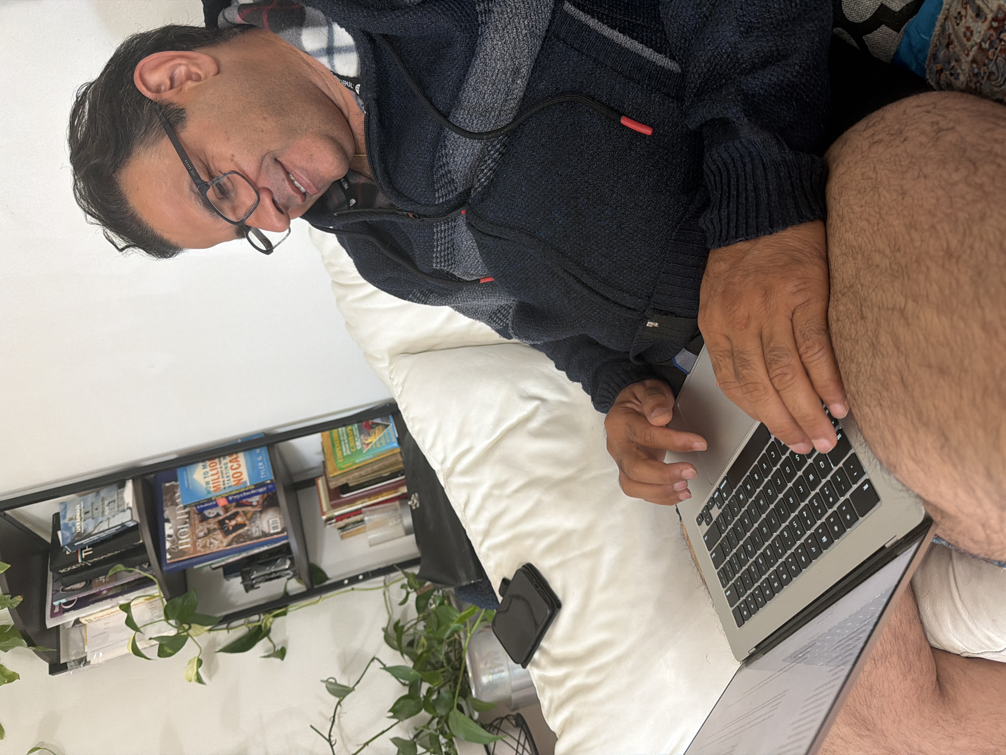 A man with dark hair and glasses sits on a white sofa, smiling and looking down at a laptop. He is wearing a dark blue hooded sweater with gray stripes. A phone is placed on the sofa next to him. In the background, a black shelf unit is filled with books and a green plant. The room appears to be a residential indoor setting.