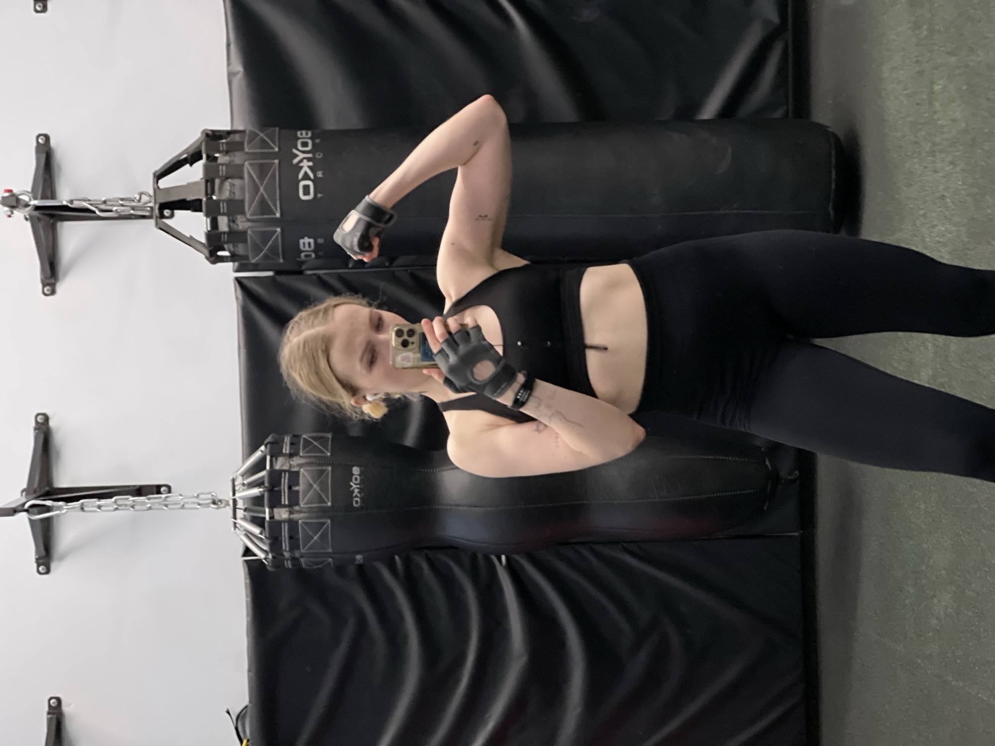 A woman is in a gym taking a selfie. She is wearing a black sports bra, black leggings, and black fingerless gloves. She is standing in front of two black punching bags suspended from the ceiling. The punching bags have the word "BOXKO" printed on them. The floor is gray. The wall is white. There is no evidence to support that the location is in Khimki, Russia.