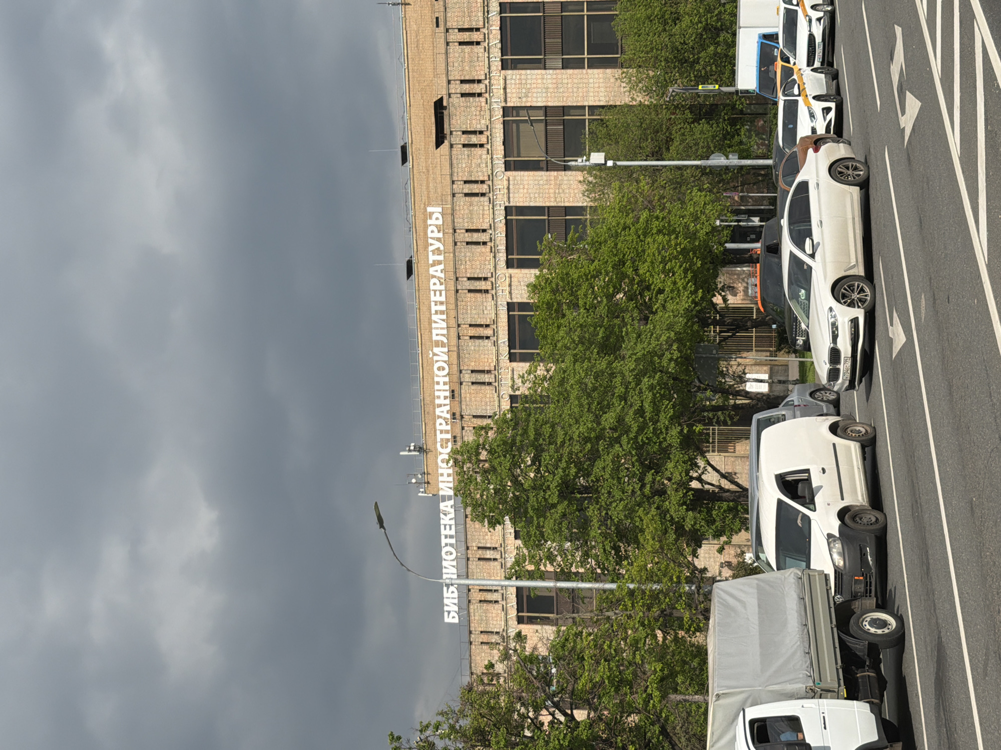 The image depicts a street scene in an unknown location, featuring a brick building with the words "БИБЛИОТЕКА ИНОСТРАННОЙ ЛИТЕРАТУРЫ" (Library of Foreign Literature) written vertically on its facade. The sky is overcast with grey clouds. The street is lined with trees and various vehicles, including white vans, cars, and a truck. The vehicles are parked along the side of the road, which has white directional arrows painted on the asphalt.
