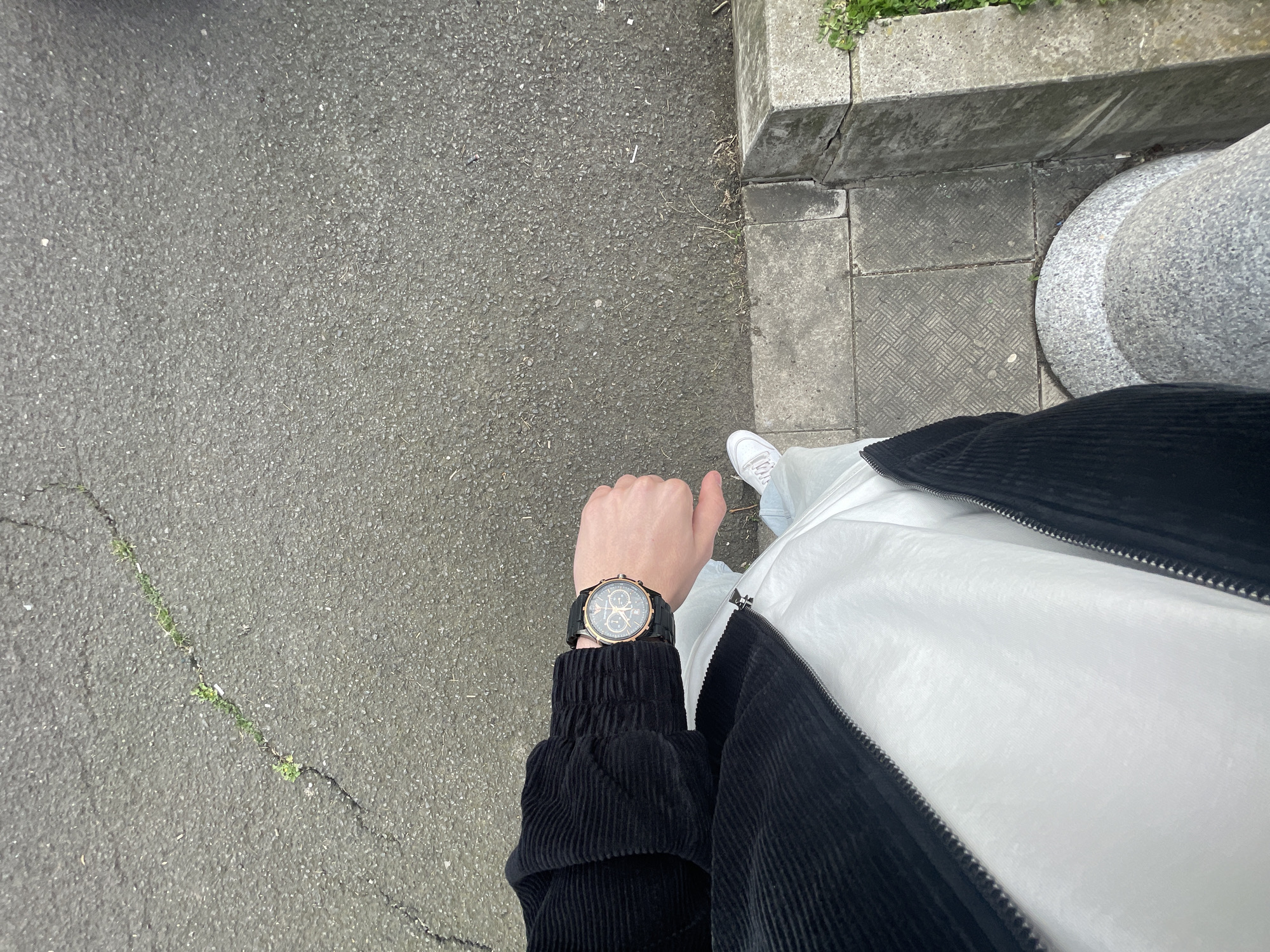 The image shows a first-person perspective looking down at a person standing on a paved surface, likely a sidewalk or road. The surface is gray and appears to be asphalt or concrete, with a crack running through it, containing small sprouts of green vegetation. To the right is a curb with a stone pillar or post. The person is wearing a white shirt under a black corduroy jacket and a watch on their left wrist. The person is also wearing white shoes. There is no explicit evidence in the image to confirm it is in Burgas, Bulgaria.
