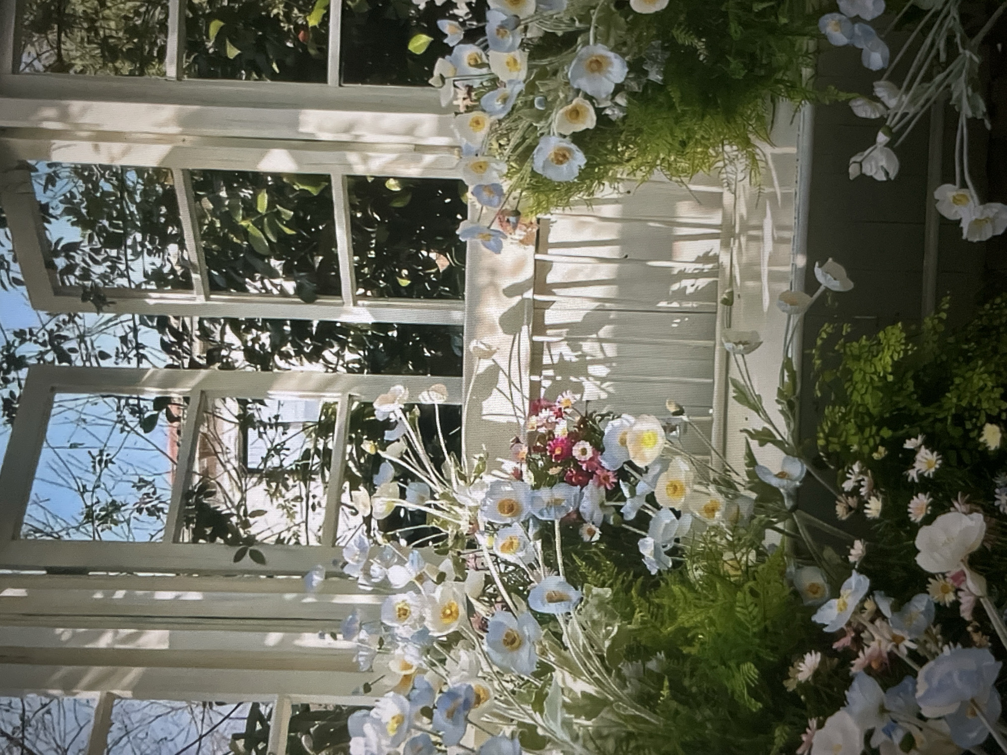 The image depicts a decorative arrangement of artificial flowers and greenery placed in front of an open white-framed window, suggesting an indoor or covered outdoor setting. The window overlooks a garden or natural landscape with trees and branches visible. The flowers are predominantly white and pale blue, with some pink accents in a smaller cluster towards the center. The overall aesthetic is light and airy, with sunlight casting shadows through the window and onto the floral display. The scene is devoid of any human presence. The location in the city of Cadek, Indonesia is not discernible from the visual information provided in the image.