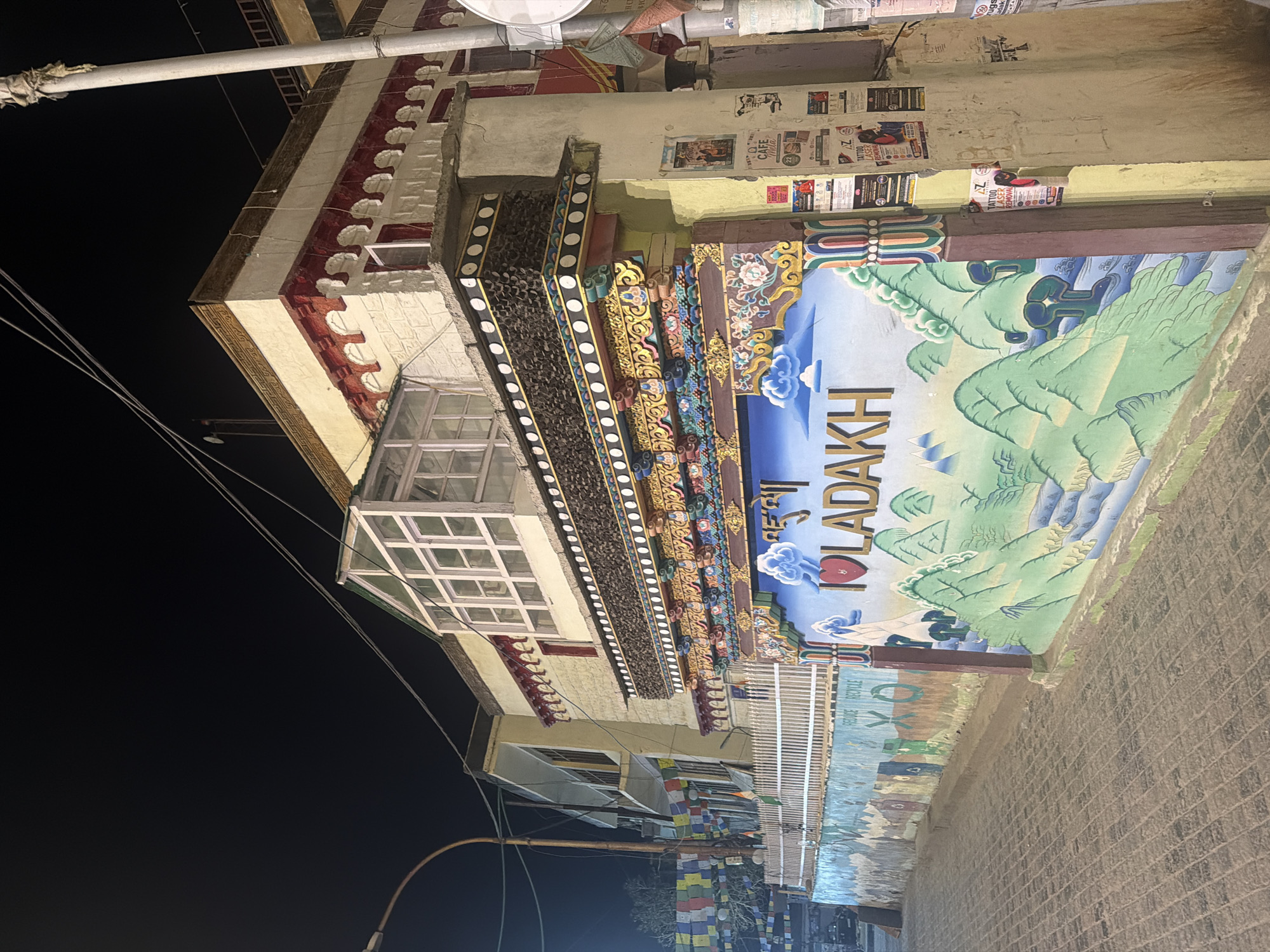 The image depicts a building facade at night in Leh, India. The building features traditional Tibetan architectural elements, including decorative eaves and colorful murals. A prominent mural on the right side of the image displays "I ❤️ LADAKH" in large letters, set against a backdrop of stylized mountains and a river. The building is illuminated by artificial light, casting shadows on the cobblestone street in the foreground. Prayer flags are visible strung across a fence on the left side of the image, suggesting a proximity to a religious or cultural site. Posters are affixed to a utility pole on the right, advertising local services. The overall scene conveys a sense of local culture and tourism.