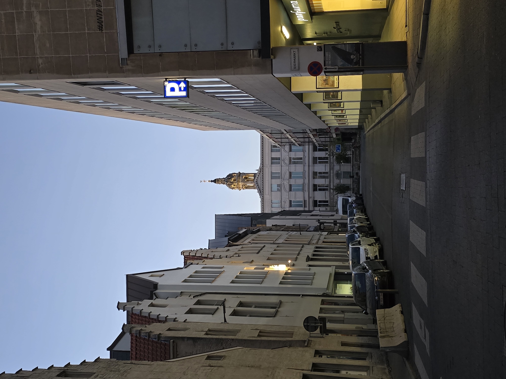 The image depicts a street scene in Antwerp, Belgium, likely during twilight. On the left, a row of historic buildings with narrow facades and distinctive gabled roofs line the street. The buildings are constructed of brick and plaster in various shades of beige and white, with some showing signs of age. In the foreground on the left, a concrete barrier is visible.
The street itself is paved with cobblestones and features a pedestrian crossing marked with white stripes. Several cars are parked along the right side of the street, creating a line of vehicles. These include a dark-colored Volkswagen Golf in the foreground, followed by a white car and several others.
The right side of the street is dominated by a modern, multi-story building with a concrete facade and a series of windows. A prominent blue parking sign with a white "P" and an arrow is mounted on the building. Below this, a sign reads "EIJERMARKT". Further down the street on the right, there is an illuminated storefront with the word "parfuma" prominently displayed. Posters are affixed to the pillars supporting the overhang of the modern building.
In the background, at the end of the street, a grand, classical-style building with a dome and spire is visible. This building is a prominent landmark and likely the Antwerp City Hall or a similar important structure. The sky above is clear and a pale blue, indicating the transition from day to night. The overall atmosphere is calm and quiet, with the streetlights and shop lights beginning to illuminate the scene.