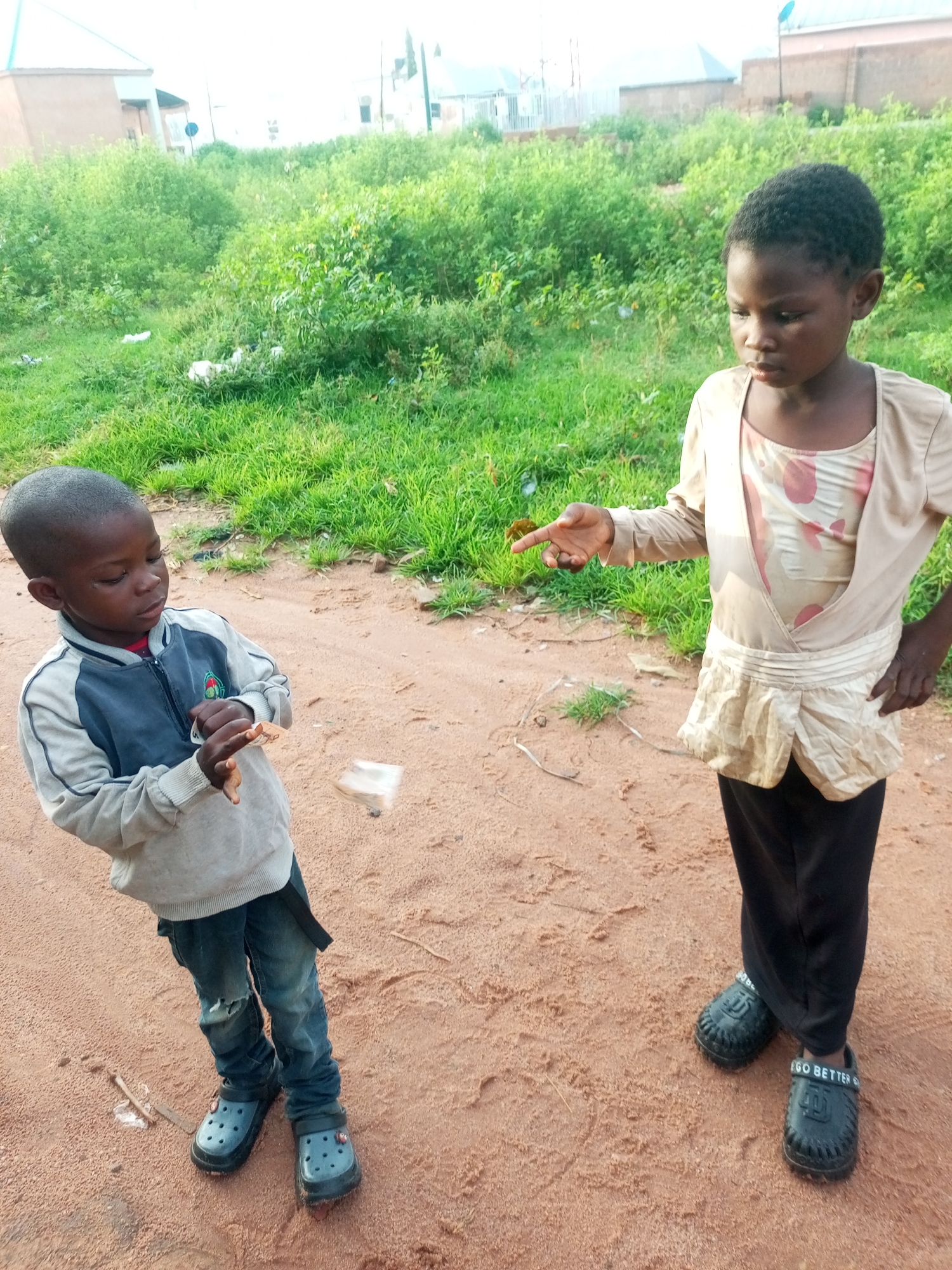 Two young children, a boy and a girl, stand in a dirt lot with sparse green grass and bushes in the background, suggesting a rural or semi-urban area within Tempure, Nigeria. The boy, on the left, is wearing a grey and blue jacket with a red shirt underneath, blue jeans with a hole in one knee, and black Croc-style shoes. He is holding a small, white object, possibly a piece of paper or fabric, in his right hand and looking down at it. To his right, the girl is dressed in a beige wrap-around top with a patterned inner layer and black pants, and she is wearing black slip-on shoes. Her right hand is extended with her index finger pointing forward, and her gaze is directed towards the boy. There is a white object suspended in mid-air between them, which appears to be the same material the boy is holding. The ground is primarily sandy dirt with some scattered twigs and debris. In the background, there are buildings and more vegetation, typical of the Tempure environment.