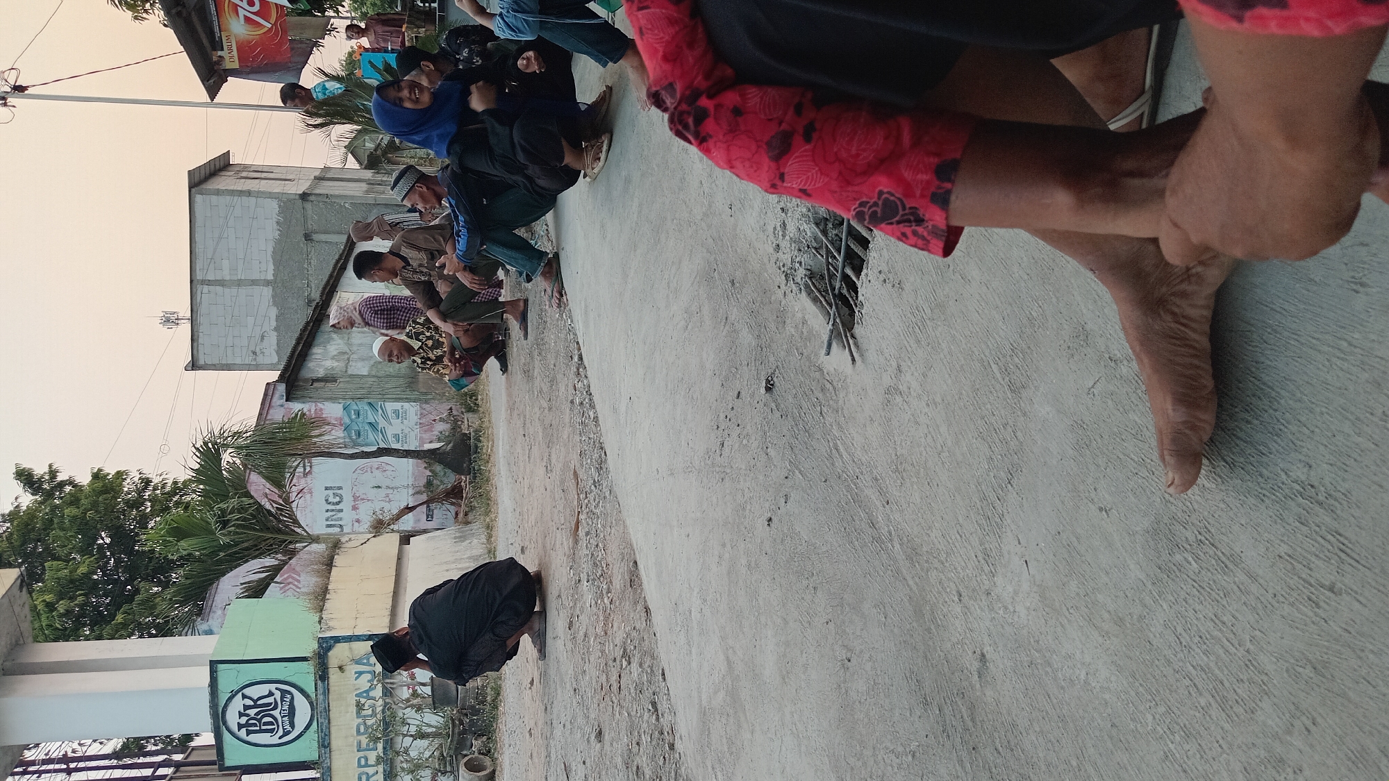 A group of people are gathered outdoors, many of them seated or squatting on the ground. The scene appears to be in Trembulrejo, Indonesia, as indicated by signage in the background. The environment is somewhat urban, with concrete surfaces and buildings visible.

In the foreground, a person's legs and feet are prominent, with one foot resting on the ground and the other crossed over. They are wearing clothing with a red and black floral pattern on the sleeves.

Further back, a man in a black cap and traditional attire is squatting on the ground, facing away from the camera. To his right, several other individuals are seated. One man wears a white skullcap and a patterned shirt, while another is dressed in a brown top. A woman in a blue hijab is also present, smiling and interacting with others.

Behind the seated individuals, there are buildings and structures. One building has a sign with "BPR JAWA TENGAH" on it, suggesting a financial institution. Other signs and graffiti are visible on the walls of the buildings. A palm tree grows in the mid-ground. The sky is a pale orange, indicating either dawn or dusk. The overall atmosphere suggests a casual gathering or waiting period.