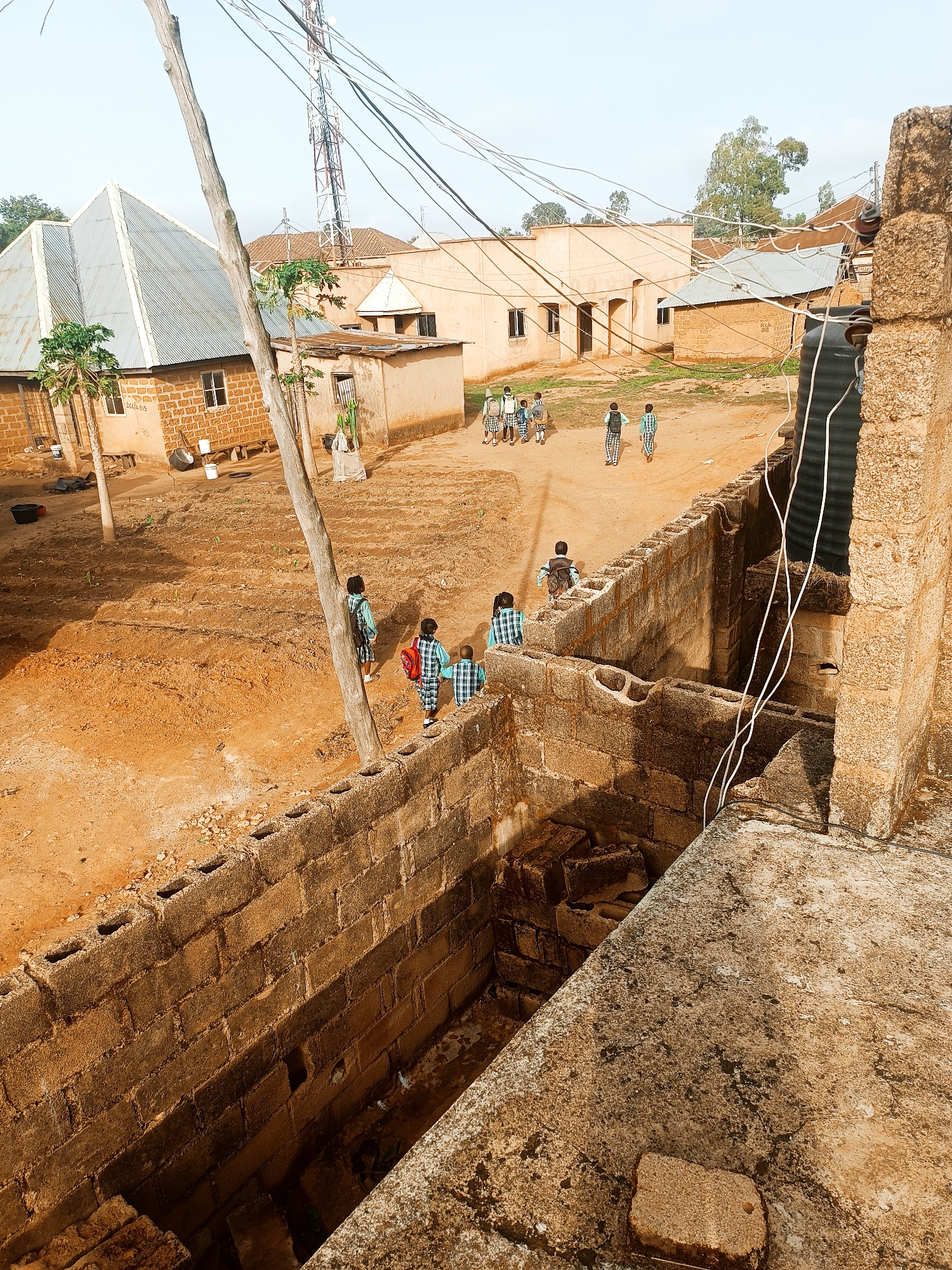 The image depicts a scene in Bukuru, Nigeria, showing a group of school children in uniform walking on a dirt path between buildings. The children are heading away from the camera. The environment consists of several buildings with corrugated metal or clay tile roofs and a cell tower in the background. The foreground features a wall made of cinder blocks. The ground is dry and appears to be dirt or reddish-brown soil. There is a mix of residential and possibly commercial buildings, suggesting a village or small town setting. Utility lines are visible overhead.