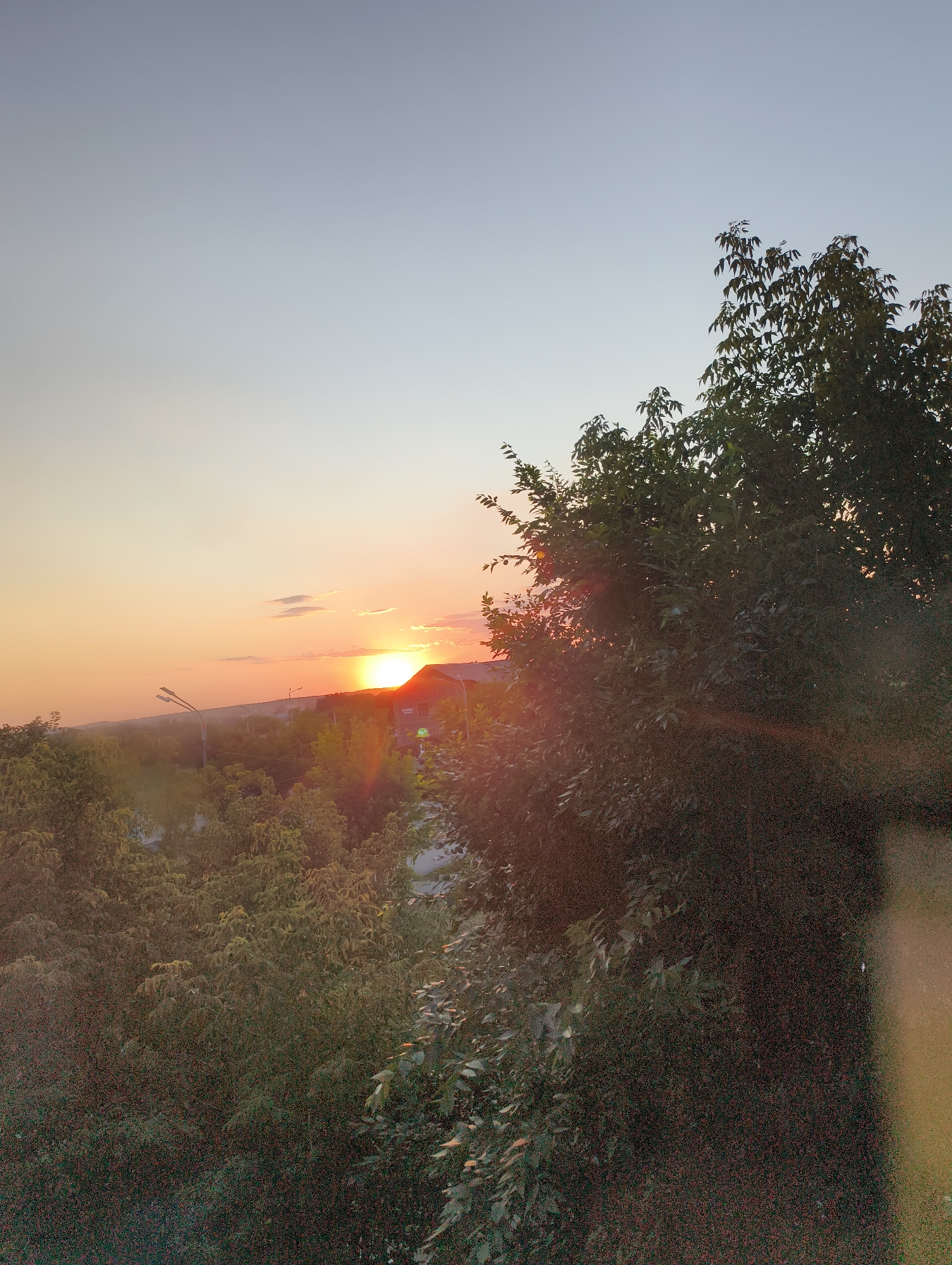 An outdoor, eye-level, vertical, medium shot captures a sunset over a tree-filled landscape in an unspecified urban location. The sky transitions from a pale blue at the top to a warm orange near the horizon, with a few wispy clouds scattered across the sky. The sun, a bright orange orb, is partially obscured by the roofline of a building, casting a warm glow on the scene.
In the foreground, the dense foliage of trees dominates the right side of the frame, with their leaves appearing dark and somewhat out of focus, likely due to the low light conditions and the lens's aperture. A subtle lens flare, a reddish-orange streak, emanates from the area of the sun and extends towards the trees.
In the mid-ground, a gently rolling landscape is visible, dotted with more trees and some structures. A street lamp with two bulbs stands prominently to the left of the frame, its metal pole extending upwards. In the distance, a building with visible signage, one word appearing to be "ГАСТРОНОМ," is situated on the right side of the sunset. The building has a bright green sign that reads "24/7."
The overall impression is one of a serene, late afternoon or early evening scene, capturing the natural beauty of a sunset juxtaposed with elements of an urban environment.