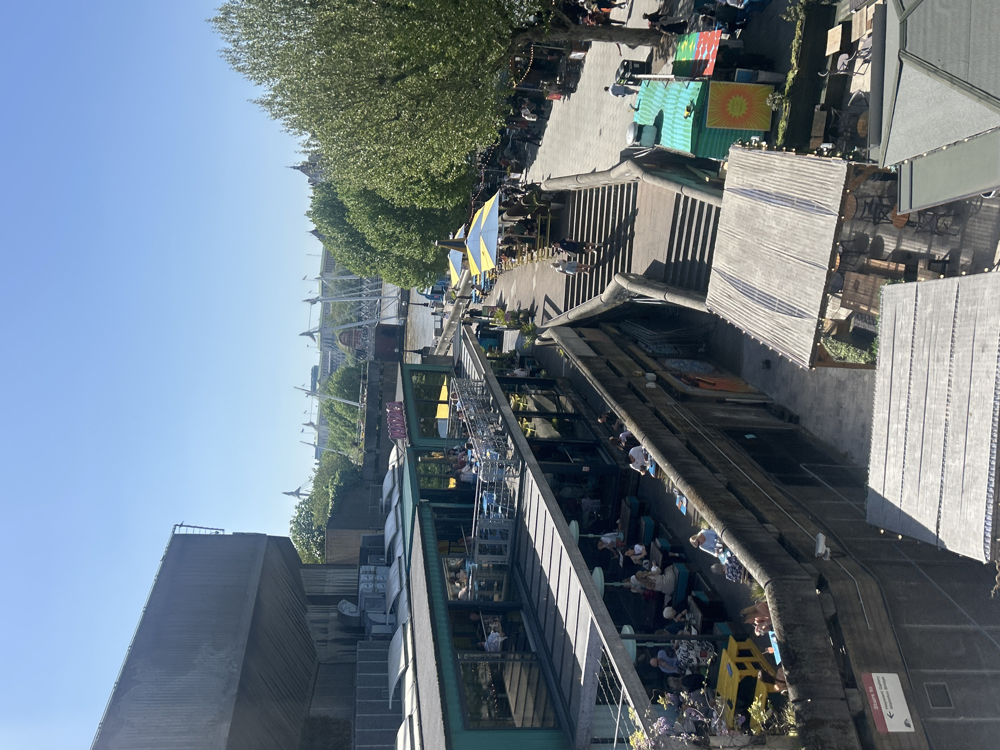 The image, which appears rotated 90 degrees clockwise, captures a vibrant outdoor scene from an elevated viewpoint, likely overlooking a bustling promenade in London, United Kingdom. The clear blue sky and bright sunshine suggest a pleasant day, likely late morning or afternoon, with long shadows visible.
The scene unfolds across multiple levels. A wide pedestrian walkway is lined with numerous mature green trees, particularly on the upper right side (when viewing the image upright). Along this promenade, many people are walking and congregating. Several outdoor dining areas are set up with tables and chairs, some sheltered by large, distinctive yellow and blue striped umbrellas. Colorful market stalls or kiosks, including one turquoise structure with a bright sun motif and another with geometric patterns, add to the lively atmosphere.
Below the main promenade, a long building with large windows and a green roof hosts what appears to be a cafe or restaurant, with additional outdoor seating areas bustling with patrons. To the far left (when viewed upright), a large, angular grey building dominates the foreground. In the distance, various urban structures, including a bridge and buildings with spires, are visible.
A brown sign mounted on a concrete barrier provides a clear location cue, with partially visible text indicating "Stairs to" with an arrow, and "Hayward Gallery Waterloo Bridge". The overall impression is one of a popular, active public space, possibly a market or leisure area along the South Bank, with people enjoying the outdoor environment.