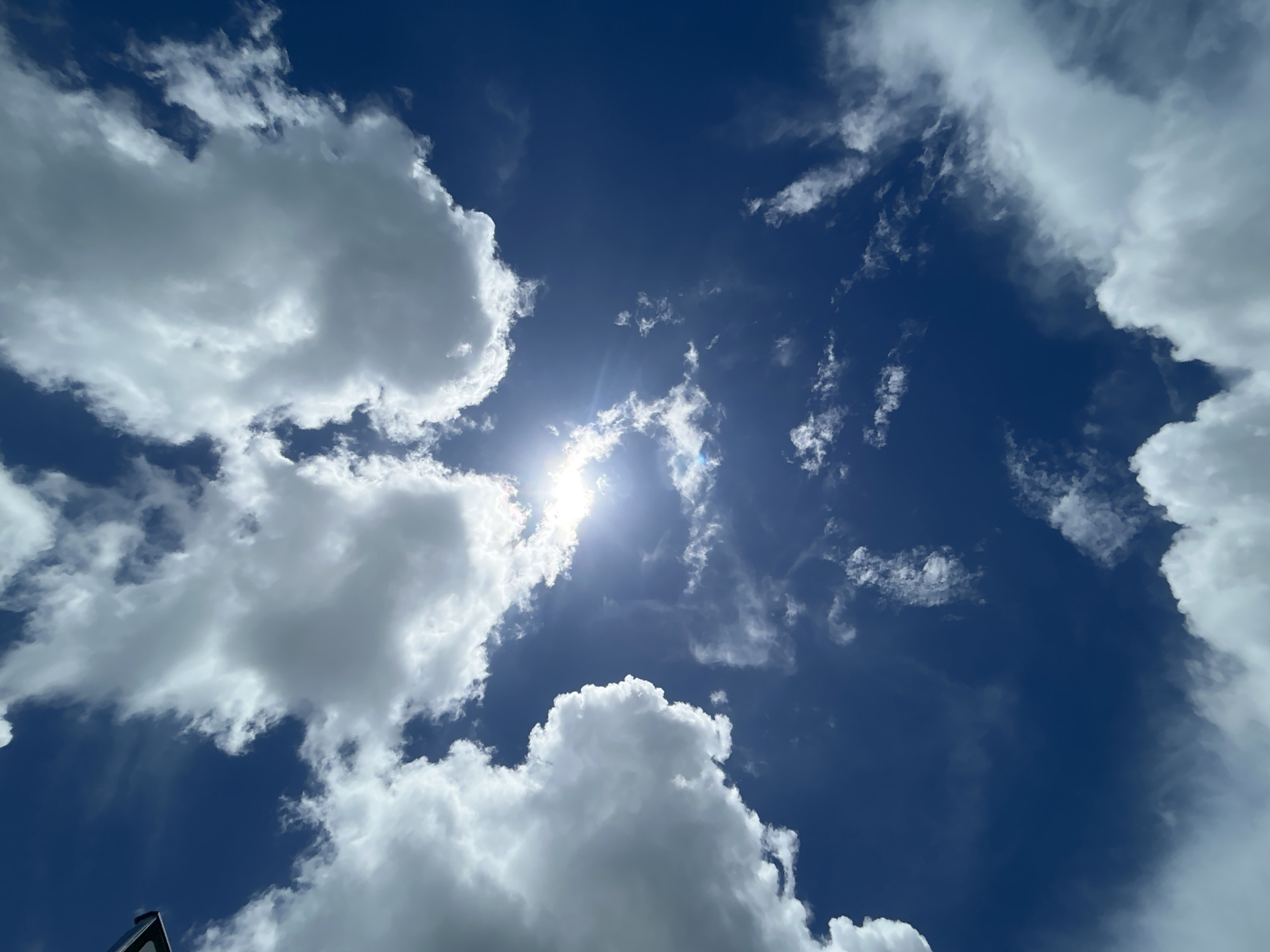 The image captures a daytime view of the sky above Baan Hin Kong, Thailand. Predominantly blue, the sky is interspersed with white, fluffy cumulus clouds of varying sizes and shapes. The sun shines brightly, partially obscured by a thin layer of cloud, creating a bright spot in the center of the image. The clouds are scattered throughout the frame, with larger clusters positioned on the left and right sides. A small, dark object is visible in the lower left corner of the image.