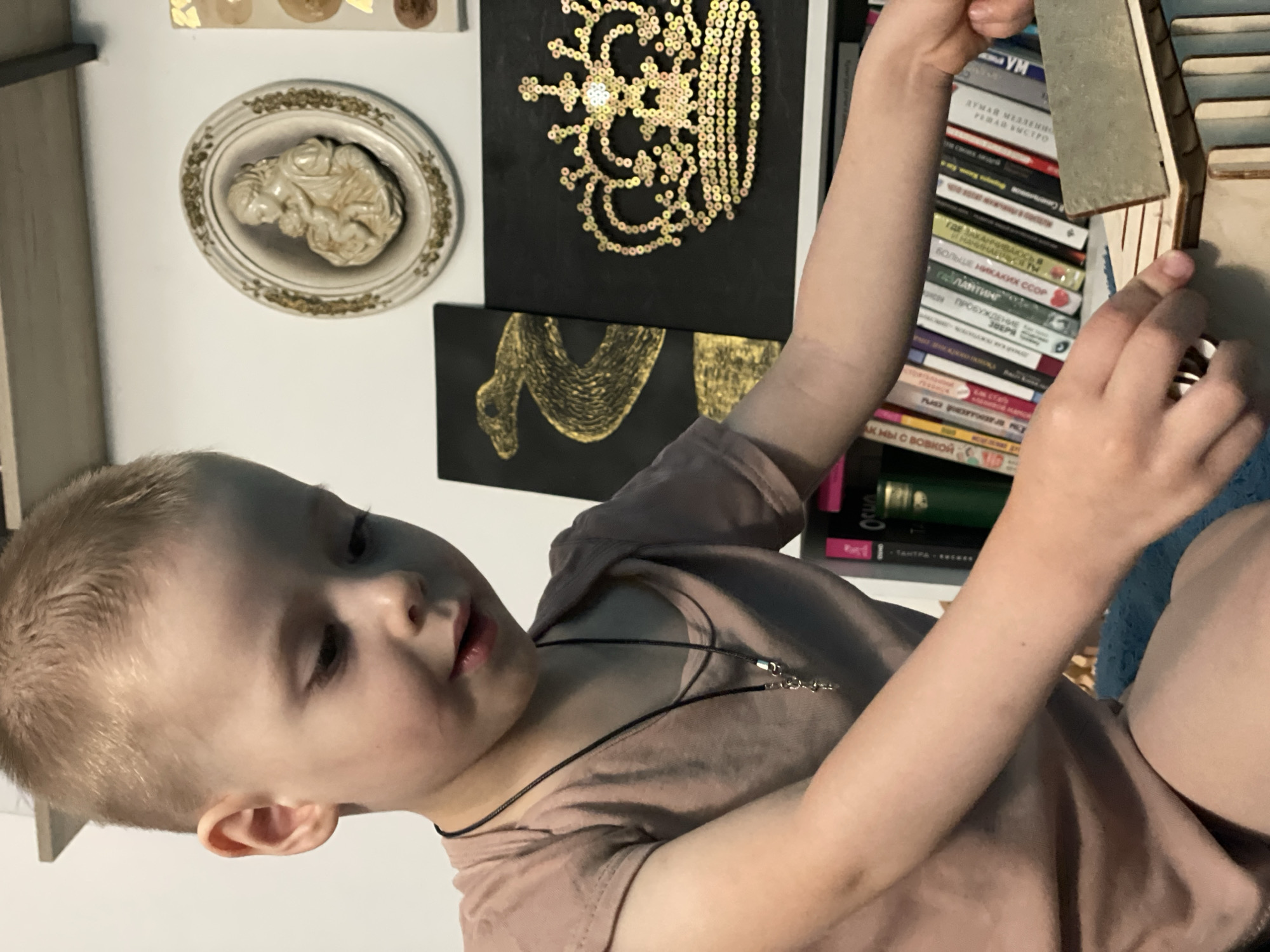 A child with fair hair and wearing a light brown t-shirt is holding a small wooden object. The child wears a black cord necklace with silver hardware. In the background, there is a white wall adorned with decorative art pieces: an oval-shaped relief, a black square with a gold sequin design, and a black square with a gold paint design. To the right of the child, there is a bookshelf filled with books of various colors and languages. The child is sitting on a blue surface.
