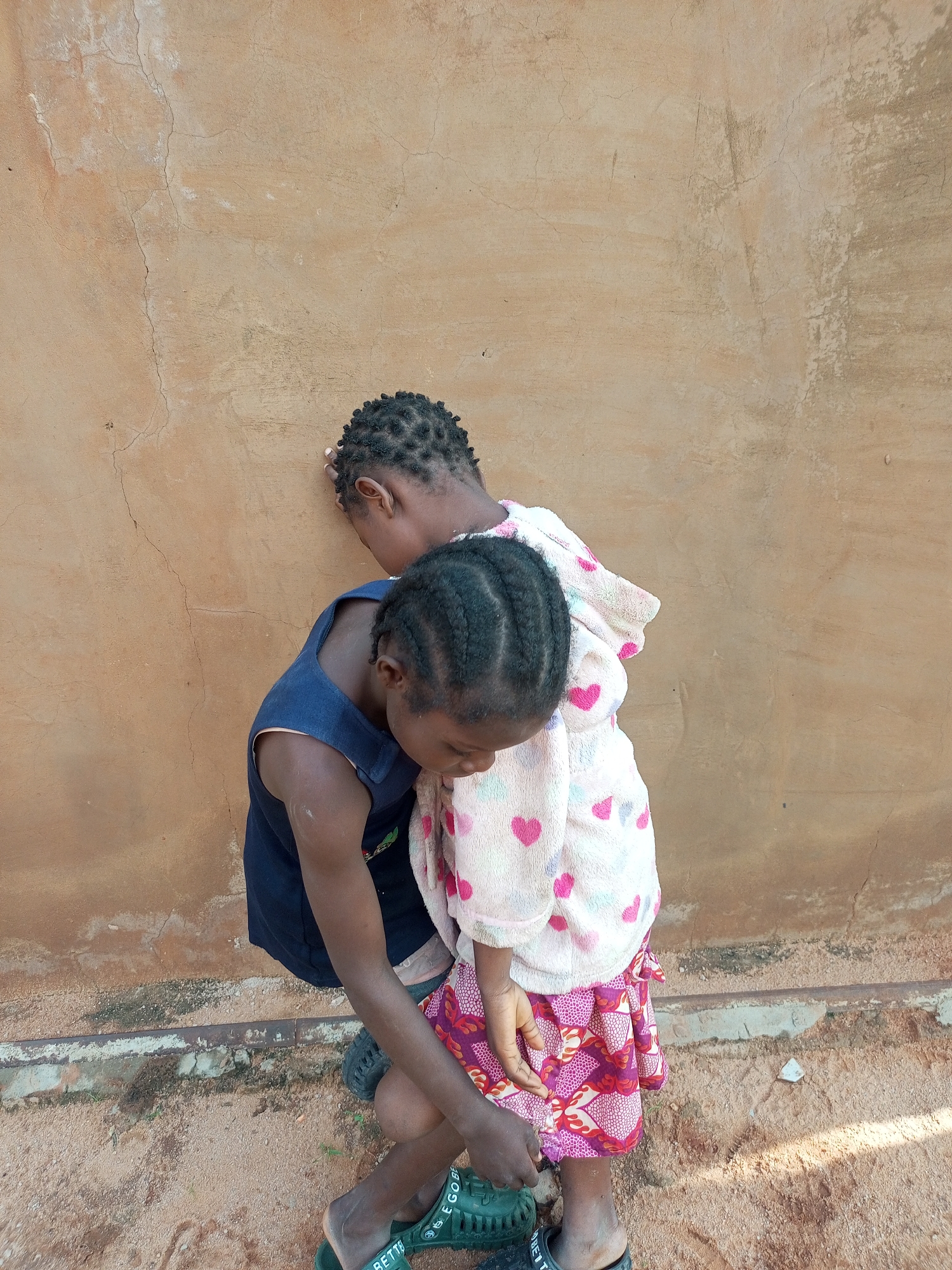 Two young girls are standing outside a building with a tan, cracked wall. The girl in front is wearing a dark blue sleeveless dress and a pink and white patterned robe with hearts. Her hair is braided, and she is looking down at her hands, which are near the skirt of the girl behind her. The girl behind her has her head turned to the side and is leaning her forehead against the wall. Her hair is also braided. Both girls are wearing sandals. The ground is sandy with some patches of dirt and small stones. The overall impression is a candid moment of two children interacting outdoors. This scene is likely located in the city of Gombe, Nigeria.