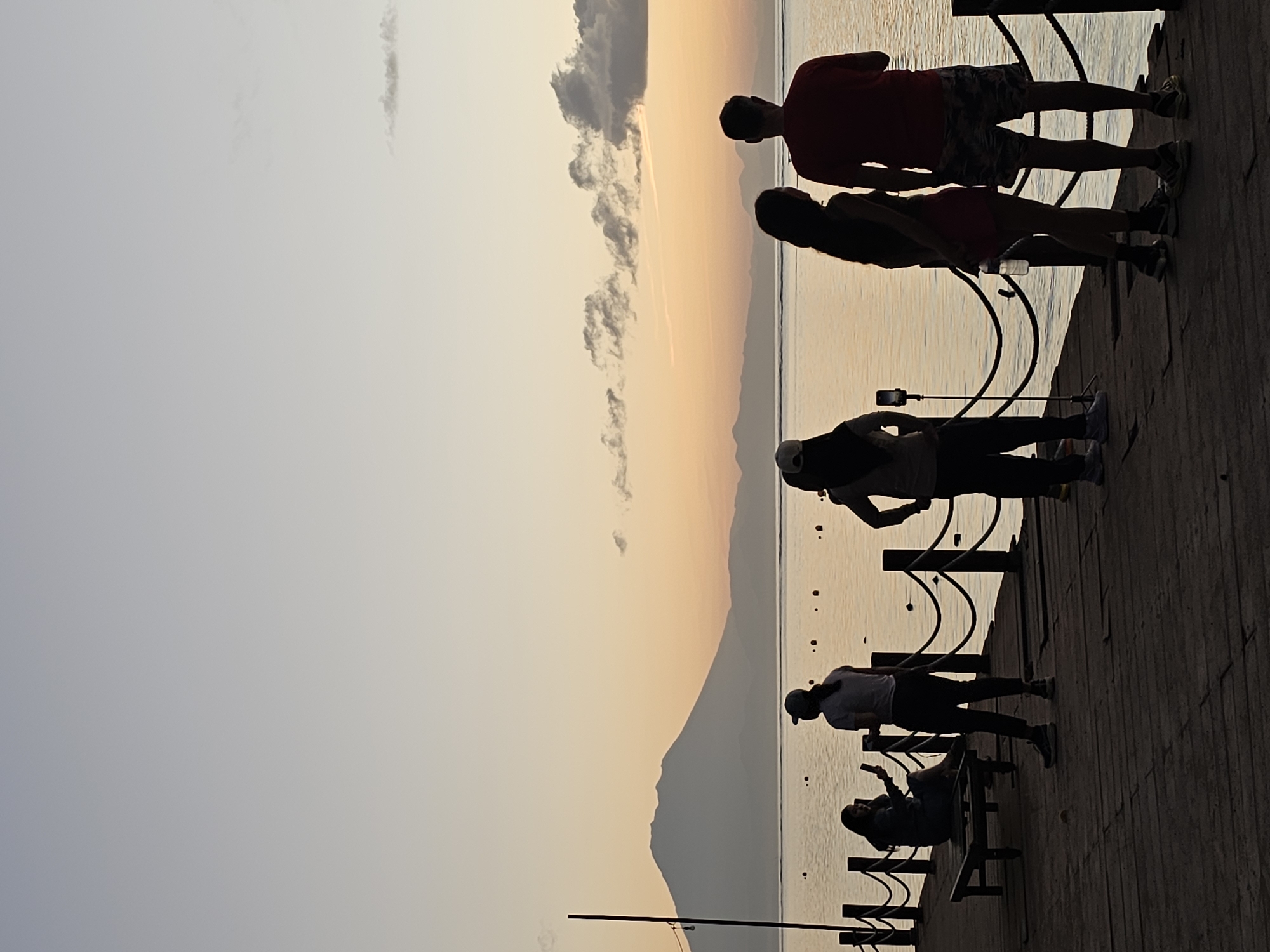 The image shows a group of people on a boardwalk overlooking the water at sunset. The location is likely Denpasar, Indonesia based on the environment.
Five people are standing or sitting on the boardwalk. From right to left:
* A man and a woman are standing close to each other, looking out at the water.
* A person is standing by a rail and holding up a mobile phone.
* A person is standing by a rail looking out to the water.
* A person is sitting on a bench looking at the water.
The boardwalk has metal railings. The background features a mountain silhouette and a sky with a gradient of colors from orange to gray. The sun is setting on the horizon.