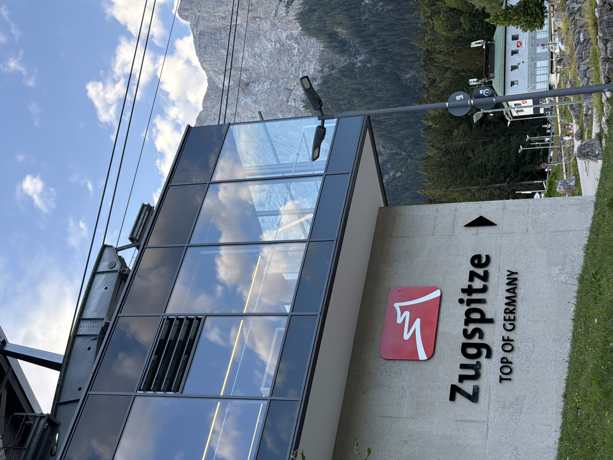 The image shows the Zugspitze cable car station in Grainau, Germany. The station is a modern building with large glass windows reflecting the sky and clouds. A prominent red sign with a white mountain graphic and the text "Zugspitze TOP OF GERMANY" is mounted on the concrete facade of the station. A tall lamppost with two lights stands in front of the building. In the background, a steep, rocky mountain covered in dark green coniferous trees rises towards a cloudy blue sky. Other smaller buildings and a road are visible in the distance. The overall scene depicts an outdoor location in a mountainous region, likely a tourist destination.