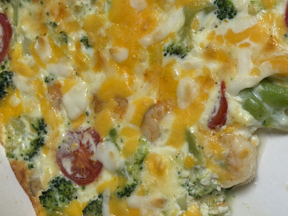 The image presents a close-up, overhead view of a colorful and hearty cooked dish, resembling a frittata, quiche, or an egg bake, served on a white plate. The main body of the dish consists of a creamy, off-white base, likely made from eggs or a dairy mixture, generously interspersed with various ingredients.
A prominent feature is the melted, gooey layer of yellow and orange cheese, which covers much of the surface and is integrated throughout the dish, suggesting it has been baked or broiled to achieve this melted texture.
Embedded within the dish are several distinct ingredients:
* Numerous bright green florets of cooked broccoli are scattered throughout, adding a vibrant color and texture.
* Multiple cooked shrimp, identifiable by their characteristic curled shape and pinkish-orange hue, are visible.
* Slices or halves of red tomatoes, possibly cherry or grape tomatoes, provide contrasting bursts of color against the yellow and green.
* There are also irregular, opaque white chunks mixed into the dish, which appear to be a type of soft cheese, such as feta or cottage cheese, or possibly larger pieces of cooked egg white.
The combination of eggs (or similar base), melted cheese, broccoli, shrimp, and tomatoes creates a visually appealing and wholesome meal. The textures appear varied, from the smooth, melted cheese to the firmer pieces of vegetables and seafood. A portion of a white plate is visible along the bottom-left and right edges of the image, indicating the dish is ready to be eaten. No text or specific environmental details like time of day or location are discernible from the image.