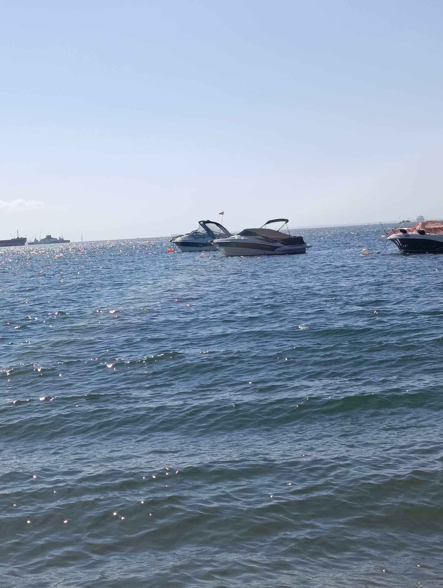The image shows a body of water, likely the Sado River estuary, with several boats. In the foreground, the water is choppy with sunlight reflecting off the surface. In the middle ground, two speedboats are anchored or moored, one with a dark hull and a tan bimini top, the other with a white hull and a dark canopy. A third boat, partially obscured, is visible to the right. In the background, several larger ships and industrial structures are visible on the horizon, suggesting proximity to a port or industrial area. The sky is clear and blue. The scene is characteristic of the waterfront area in Setúbal, Portugal, particularly near the commercial port or leisure marina.