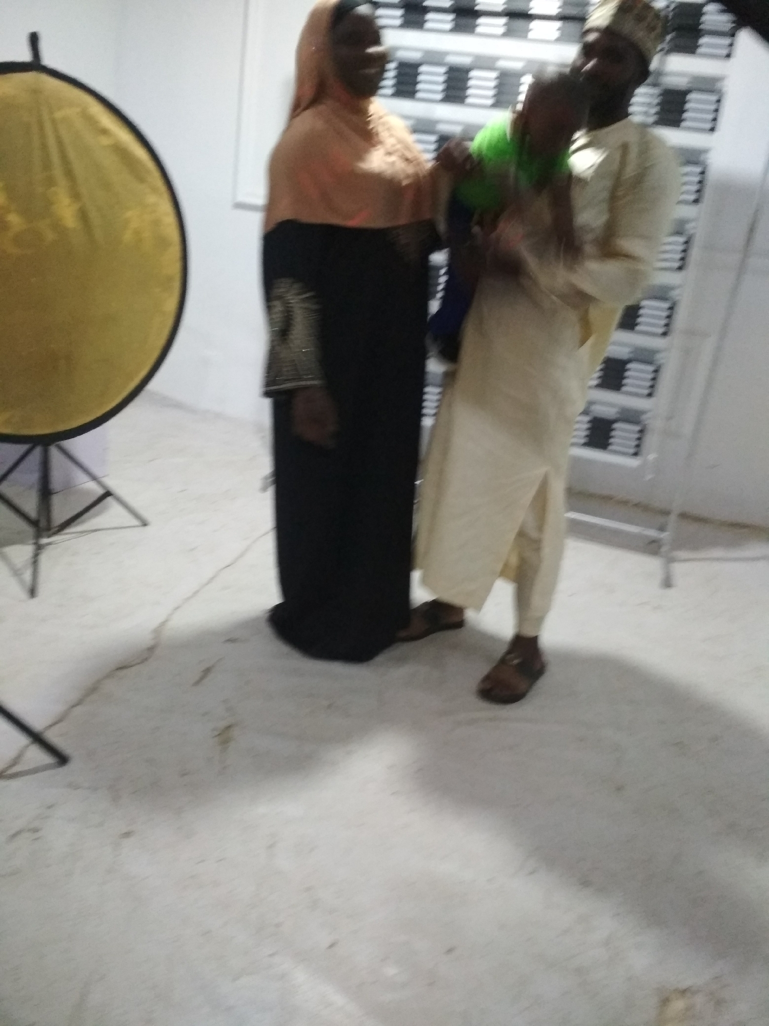 The image shows a man, a woman, and a small child in an indoor setting. The woman is wearing a long black dress with an orange hijab. The man is dressed in a cream-colored traditional robe and a matching hat, holding the child. A large gold reflector is positioned to the left of the frame. The background includes a wall with a pattern of repeating horizontal lines, possibly bookshelves, and a metal stand. The floor is light gray. The location is Unknown, Unknown.