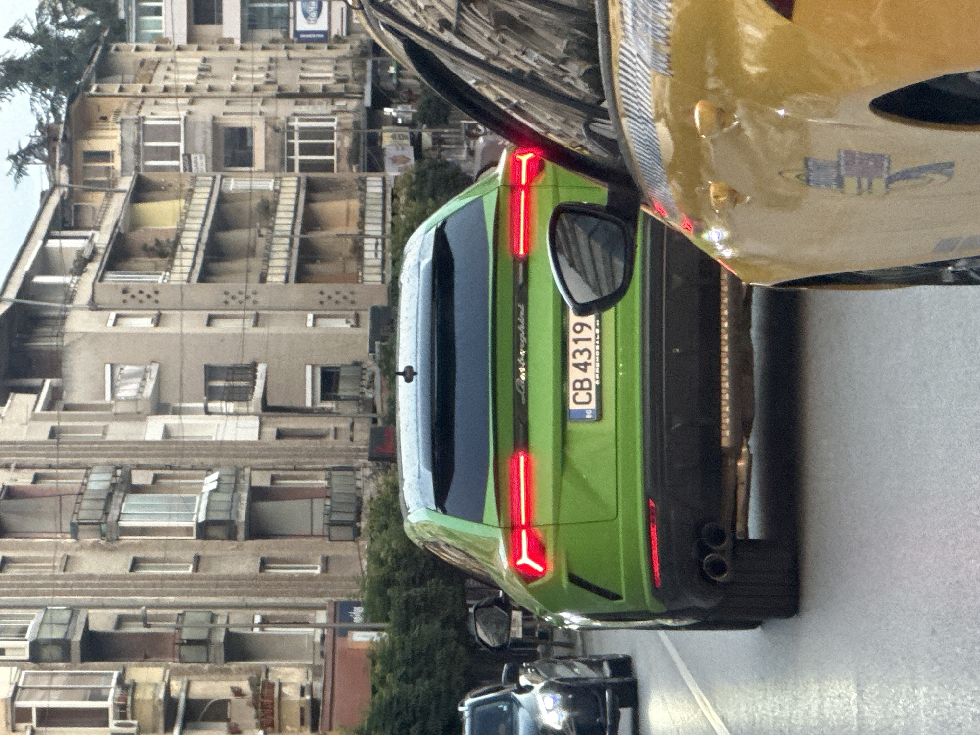 The image shows the rear of a bright green Lamborghini Huracán parked on a street in Veliko Tarnovo, Bulgaria. The car's distinctive Y-shaped taillights are illuminated. A license plate with the characters "CB 4319" is visible. The car is positioned next to a yellow vehicle, suggesting a car meet or a similar event. In the background, a multi-story building with numerous windows and balconies is visible, indicative of an urban setting. The lighting suggests it is daytime, possibly overcast. The overall scene is a snapshot of a luxury car in a residential or urban environment in Bulgaria.