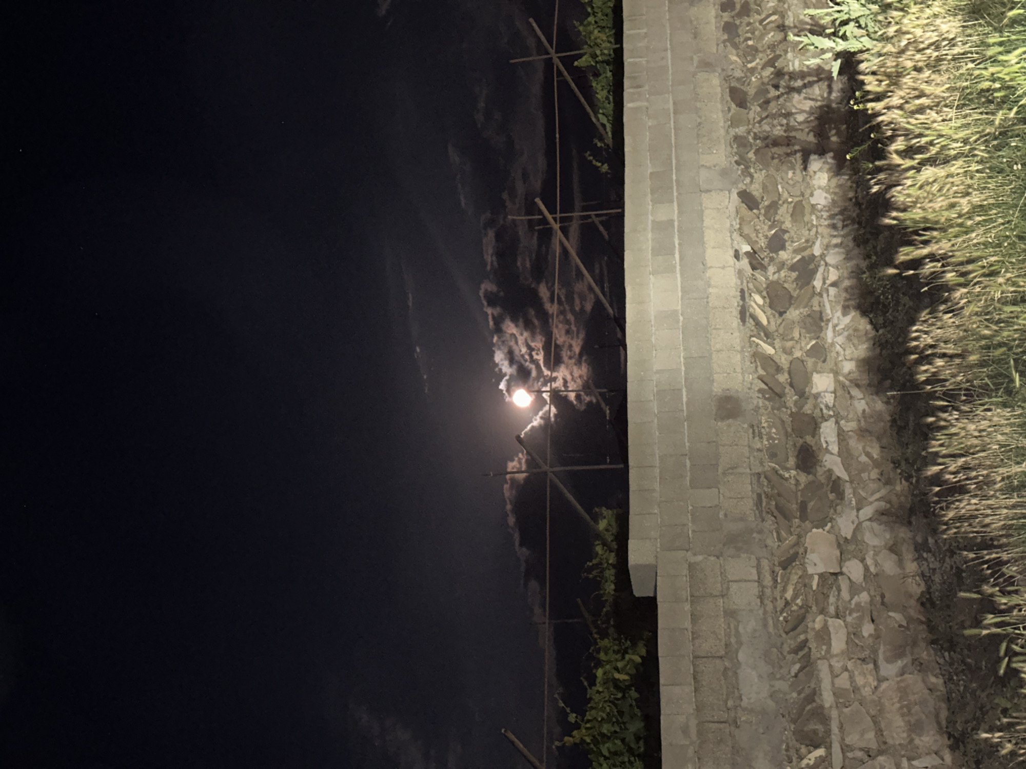 The media file shows a night scene, rotated for vertical display. The left side of the image is dominated by a dark night sky with some scattered clouds. In the center, a bright, full moon is partially obscured by clouds, emitting a soft glow. A wooden trellis or scaffolding structure is visible in front of the sky, with some green foliage clinging to it.
To the right of the sky, a stone wall is constructed from rectangular blocks, leading down to a section of rougher, natural stone paving. The stone wall is illuminated, casting shadows on the ground. To the far right, tall, dry grass or reeds are illuminated by a light source, appearing golden in the light. The overall impression is a serene, possibly rural or garden setting at night, with the moon as the primary light source in the sky. The location context suggests this scene is in Subutli-Miatli, Russia.