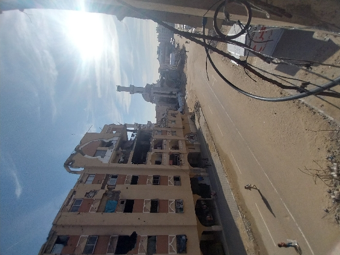 A damaged multi-story building stands alongside a road, with a mosque visible in the background. The building's upper floors are severely affected, with broken walls and missing sections. The street is largely empty. Power lines crisscross above the road. The overall scene conveys destruction and abandonment. The sun shines brightly, adding contrast to the devastation.