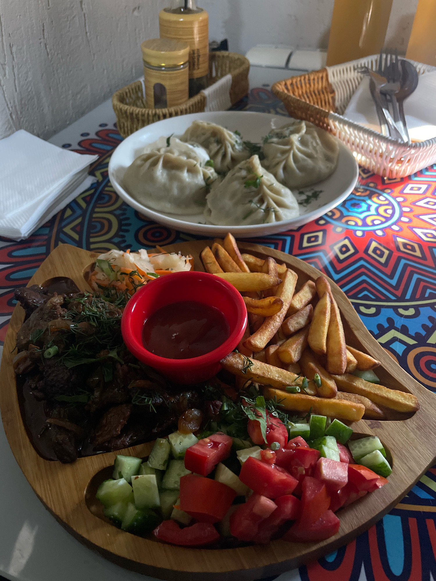 A close-up shot captures a wooden platter filled with food on a table with a colorful, patterned tablecloth. The platter is divided into sections. One section contains a pile of dark, braised meat topped with fresh green herbs. Next to it is a small red bowl filled with a dark red sauce, likely ketchup. Adjacent to the sauce is a generous portion of golden french fries. Further along the platter, a fresh salad is arranged, featuring diced tomatoes and cucumbers. In the background, a white bowl holds several steamed dumplings, garnished with herbs. To the left of the platter, a stack of white napkins is visible, and to the right, a woven basket holds silverware. The table is set with condiments in jars and other dining accessories. The lighting suggests an indoor setting, possibly a restaurant.
