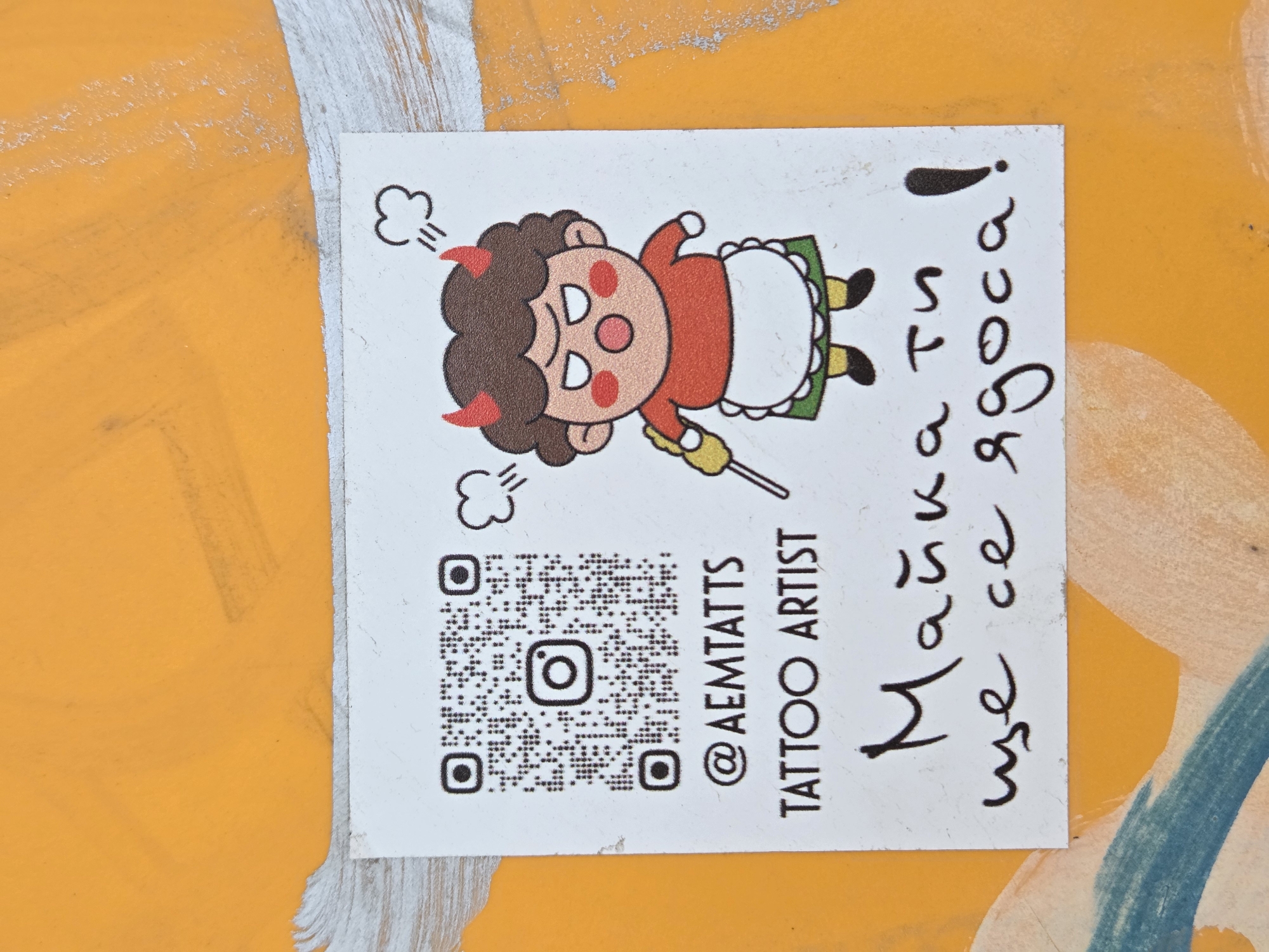 A small, rectangular sticker adhered to a yellow surface, likely an exterior wall, possibly graffiti-covered in Varna, Bulgaria. The sticker features a cartoon drawing of a devilish character, possibly a child, in a red garment, white apron, with red horns, dark hair, and a yellow object in its hand. Above the character, there are two "puff" symbols. Below the character is a QR code and the text "@AEMTATTS TATTOO ARTIST" printed in black. To the right of the image is text in Cyrillic script, also in black. The surface to which the sticker is adhered is yellow with a blue streak and some silver color. The sticker itself is white. The orientation of the sticker is turned to the side.
