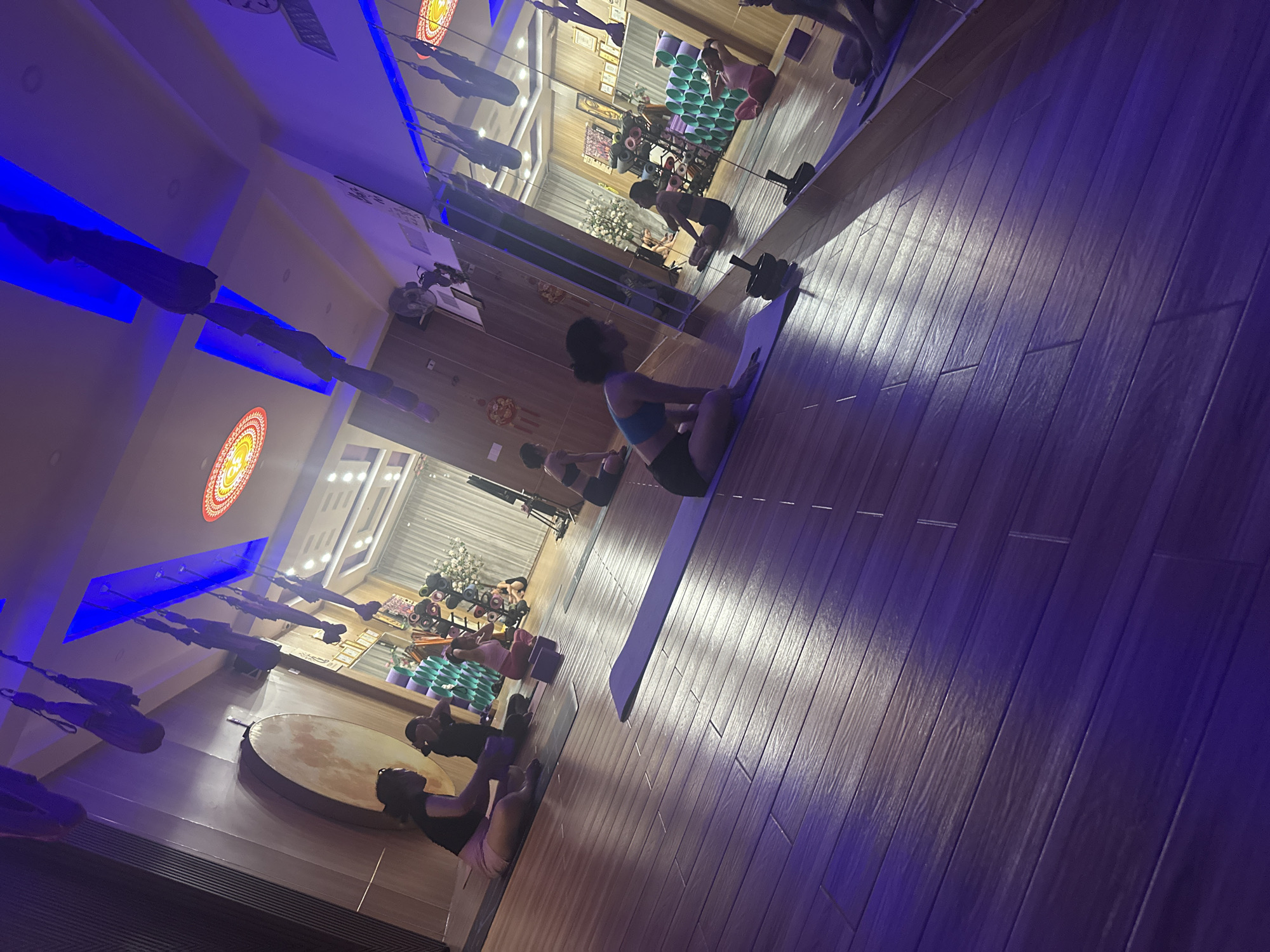 The image shows a yoga studio in Bac Nha Trang Ward, Vietnam. Several individuals are engaged in yoga poses on mats. The studio has hardwood floors and is lit with blue-toned lighting. In the background, there's a wall with mirrors reflecting the studio and participants. Yoga equipment is visible.