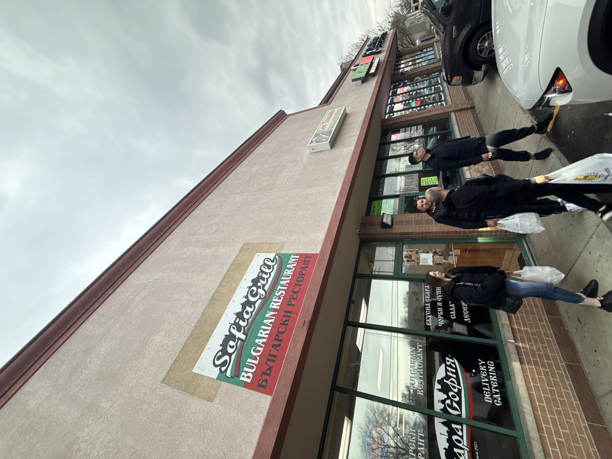 The image shows the Sofia Grill, a Bulgarian restaurant located in Des Plaines, United States of America. The restaurant has a sign in English and Bulgarian, stating "Bulgarian Restaurant" and "БЪЛГАРСКИ РЕСТОРАНТ." The establishment date is 2021. The building appears to be a strip mall with multiple storefronts. There are people walking on the sidewalk in front of the restaurant, some carrying bags. Cars are parked along the curb. The weather is overcast.