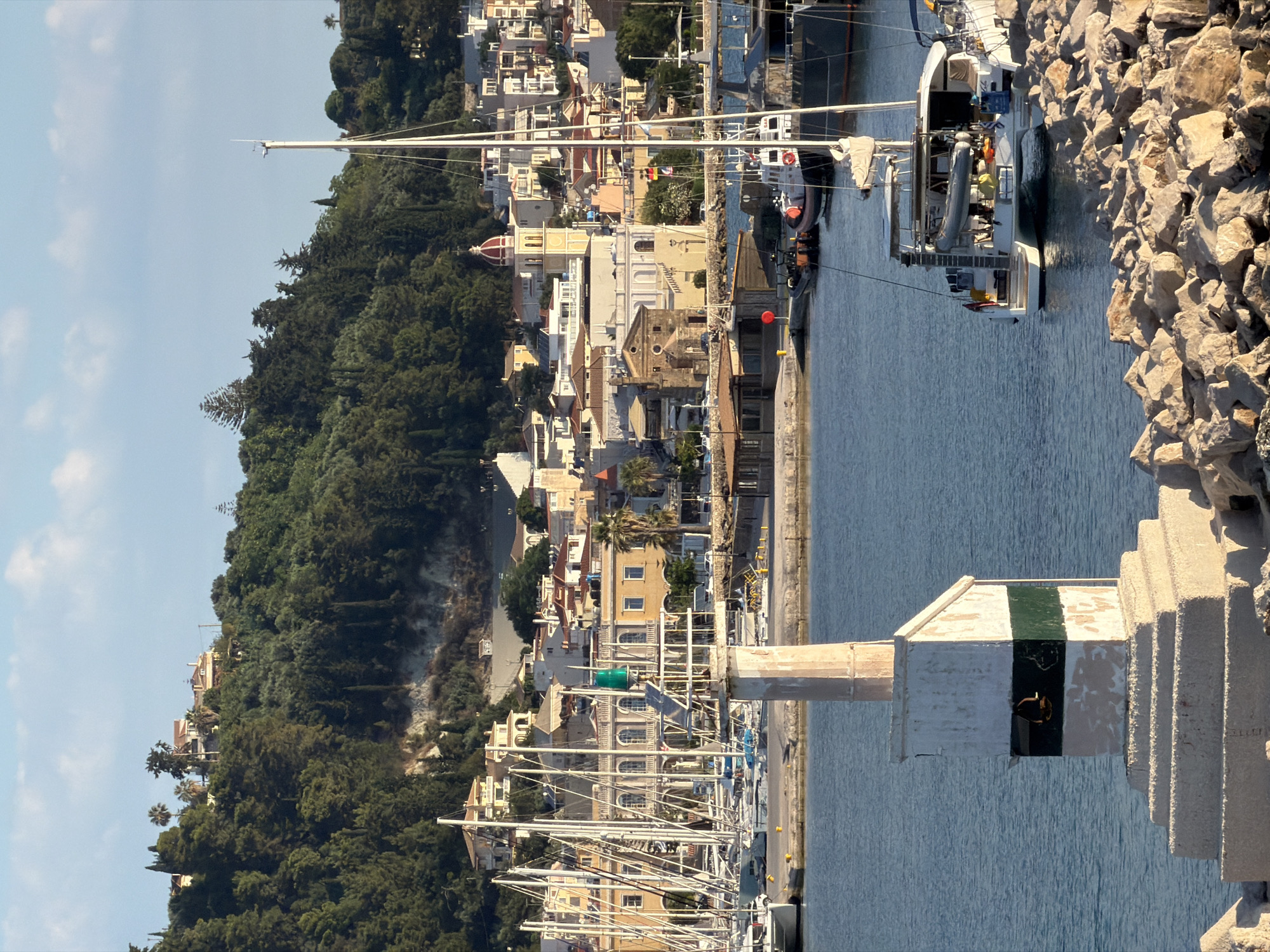 A daytime, eye-level, medium shot captures a harbor in Zakynthos Municipal Unit, Greece, with a lighthouse in the foreground. The lighthouse, made of weathered concrete and painted with white and green bands, stands on a rocky breakwater. Behind it, a bay is filled with a variety of boats, including sailboats and a catamaran docked in the foreground. In the middle ground, a picturesque town with white and yellow buildings, red-tiled roofs, and a prominent church with a dome stretches along a hillside. Lush green trees and vegetation cover the upper slopes of the hill, with a clear blue sky above. The water is calm and reflects the sunlight. There are no people visible in the scene.