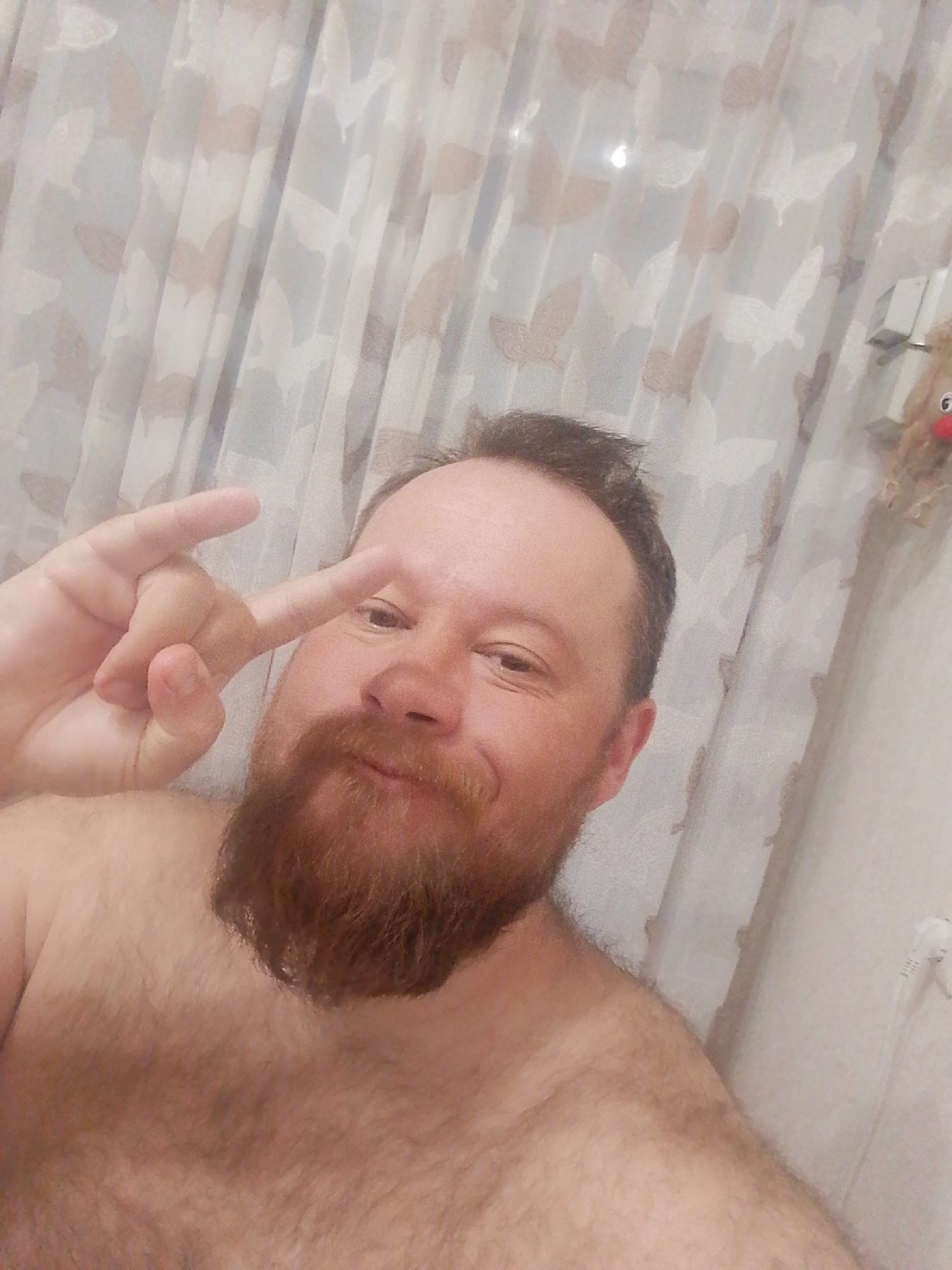 A man with a reddish beard poses shirtless in a room. He makes a gesture with his fingers near his eye. Behind him is a patterned curtain and a small reindeer decoration hanging on the wall. A power outlet is visible in the lower right. The location is unknown, and Фоки, Russia, cannot be verified.