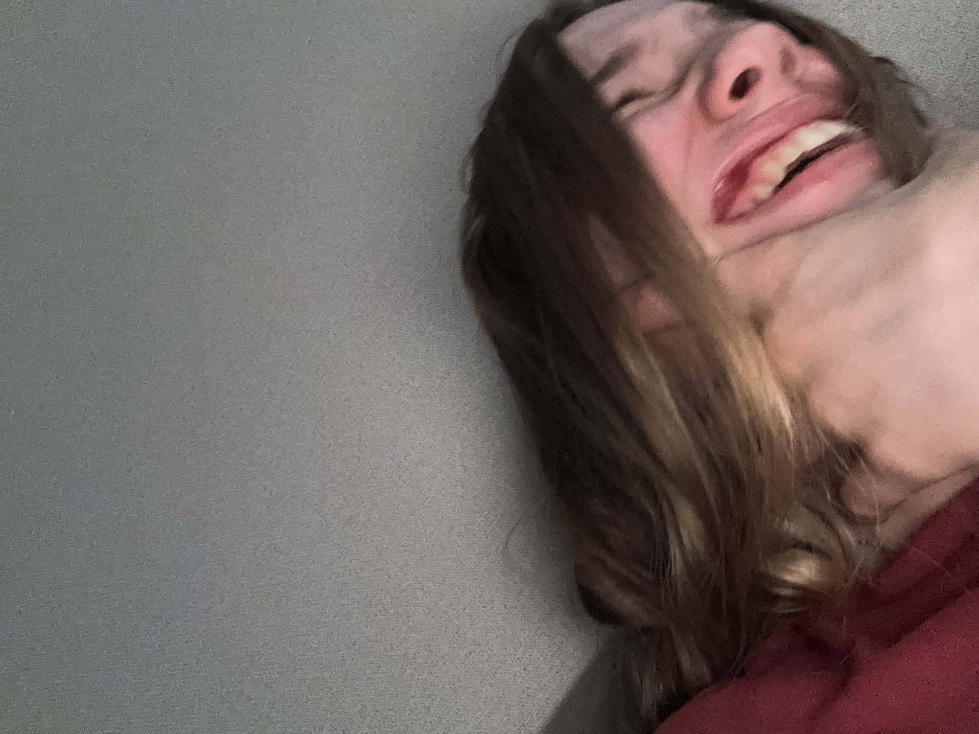The image shows a close-up of a person's face, partially obscured by their hair and a red garment. The person's mouth is open, revealing teeth. The background is a textured, gray surface. There is no discernible location within the city of Vsevolozhsk, Russia.