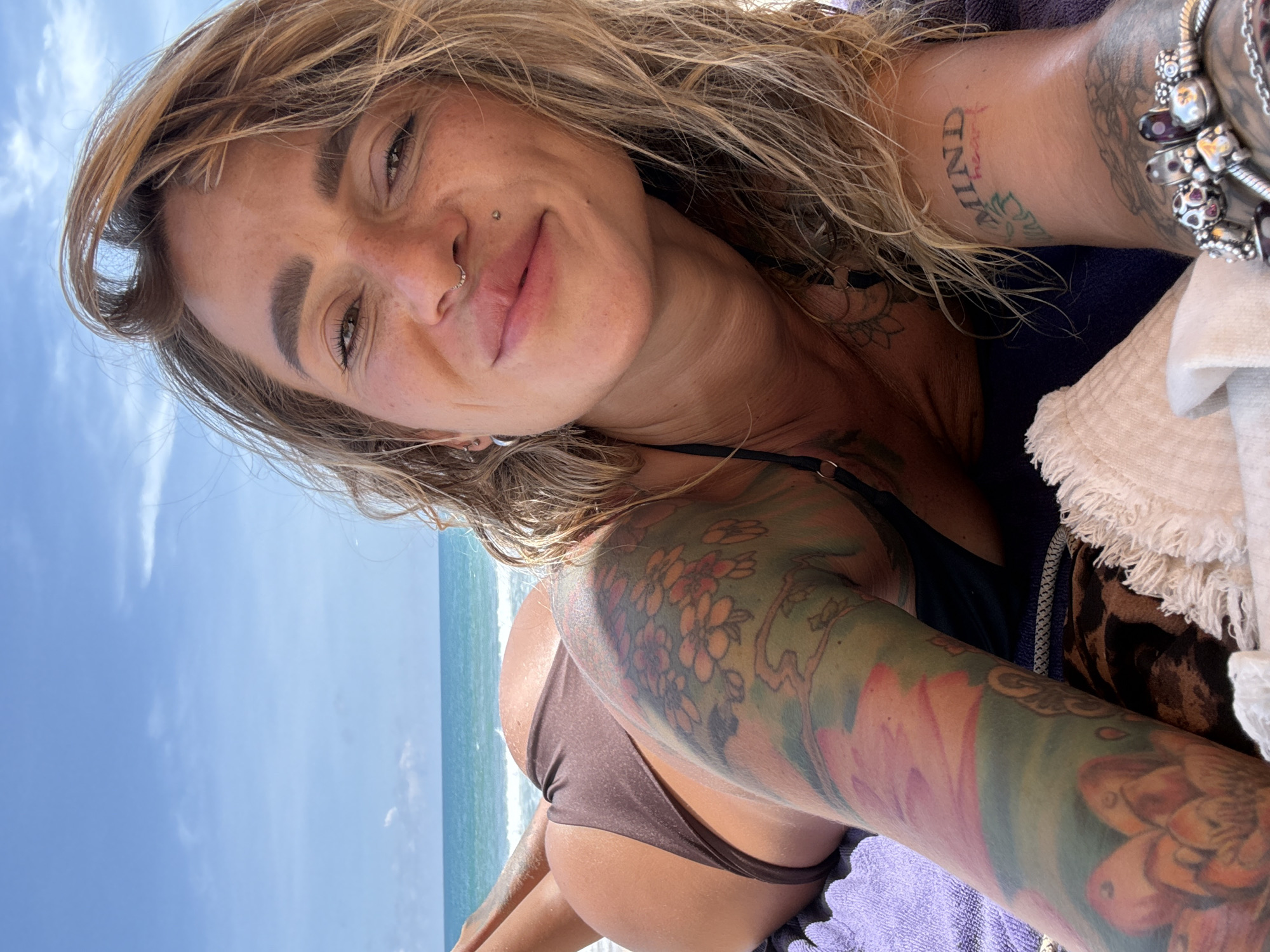 A woman with blonde hair and tattoos lies on a beach in Choeng Thale, Thailand, facing the camera with a slight smile. She wears a brown bikini bottom and a black bikini top. The ocean and sky are visible in the background. A purple towel and a tan cloth are placed beneath her.
