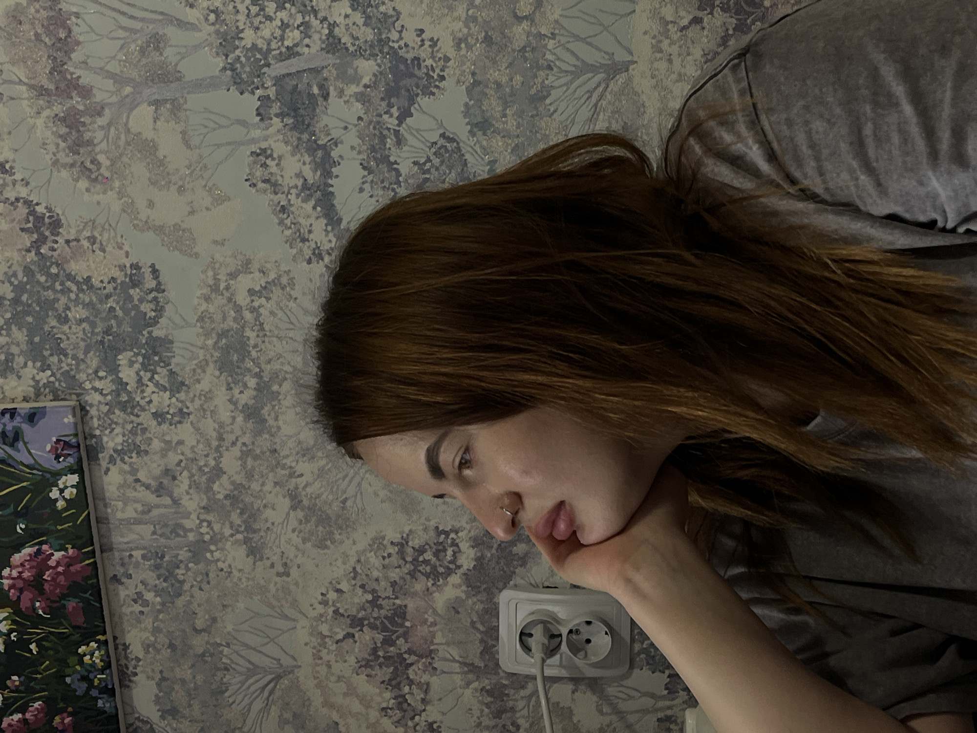 A person with shoulder-length auburn hair is resting their face on their hand, appearing contemplative. The person has fair skin and a nose piercing. They are wearing a gray t-shirt. Behind the person is a wall with a floral wallpaper pattern in shades of white, gray, and purple. A white electrical outlet with a plug inserted is visible on the wall. A framed painting with flowers is partially visible on the left side of the image. There is no information given to determine the specific location in the city of Novopokrovskaya, Russia.