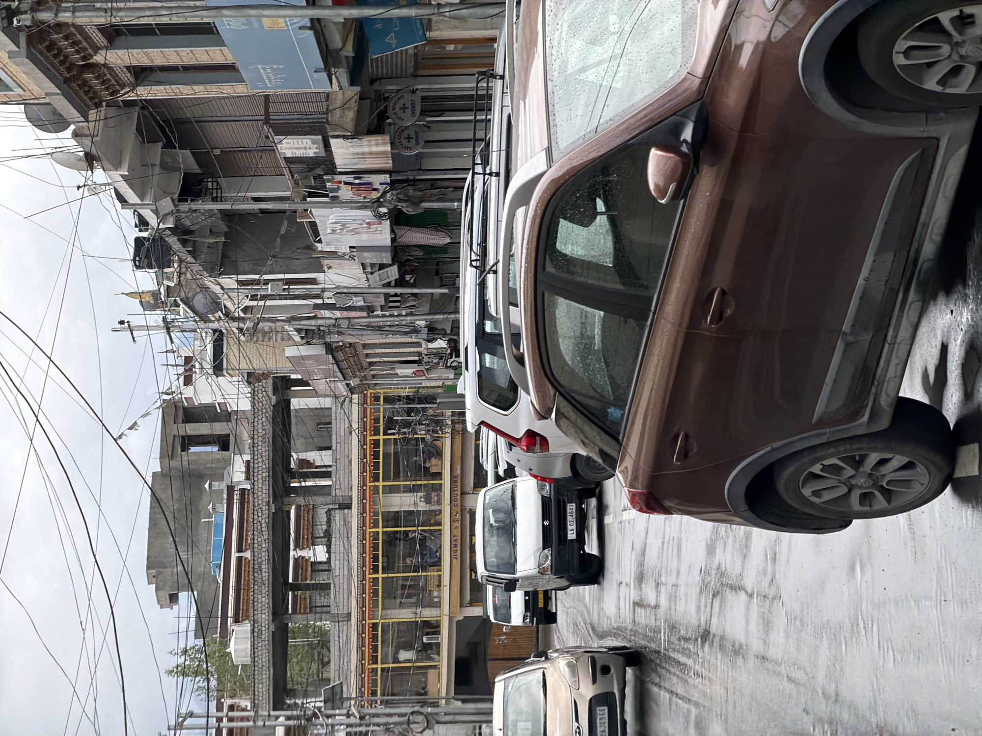 The image depicts a street scene in Leh, India, characterized by a line of vehicles on a wet road under an overcast sky. Several cars are visible, including a brown Hyundai i20 in the foreground, a white Maruti Suzuki Alto in the middle distance, and other vehicles further down the street. The buildings lining the street are multi-story structures with shops on the ground floor. One shop is identified by signage as "JIGMAT COUTURE". The architecture features elements common in Leh, with exposed brick and concrete structures, and numerous electrical wires crisscross overhead. The wetness of the road suggests recent rainfall. No people are clearly discernible within the vehicles or on the street. The overall impression is a somewhat cluttered urban environment in cloudy weather.