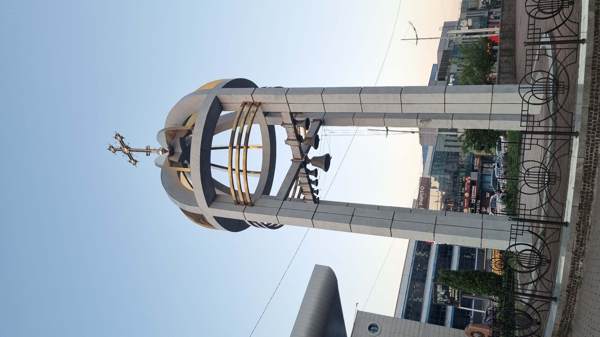 This is a low-angle, vertical photograph of an outdoor sculpture in Kyiv, Ukraine. The sculpture is made of light-colored concrete pillars and features a large, circular, crown-like structure adorned with gold accents. Suspended from the sculpture are several bells. A cross is visible at the very top of the sculpture, suggesting a religious or memorial purpose.
The setting appears to be an urban environment, with modern buildings visible in the background. One building has prominent signage including "PAPITO" and what appears to be other commercial text in Cyrillic. A decorative metal fence with circular motifs is in the foreground. The sky is clear and light blue, indicating it is daytime, likely late afternoon or early morning due to the soft lighting. No people are visible in the frame. The overall mood is calm and serene, focusing on the architectural and artistic elements of the sculpture.