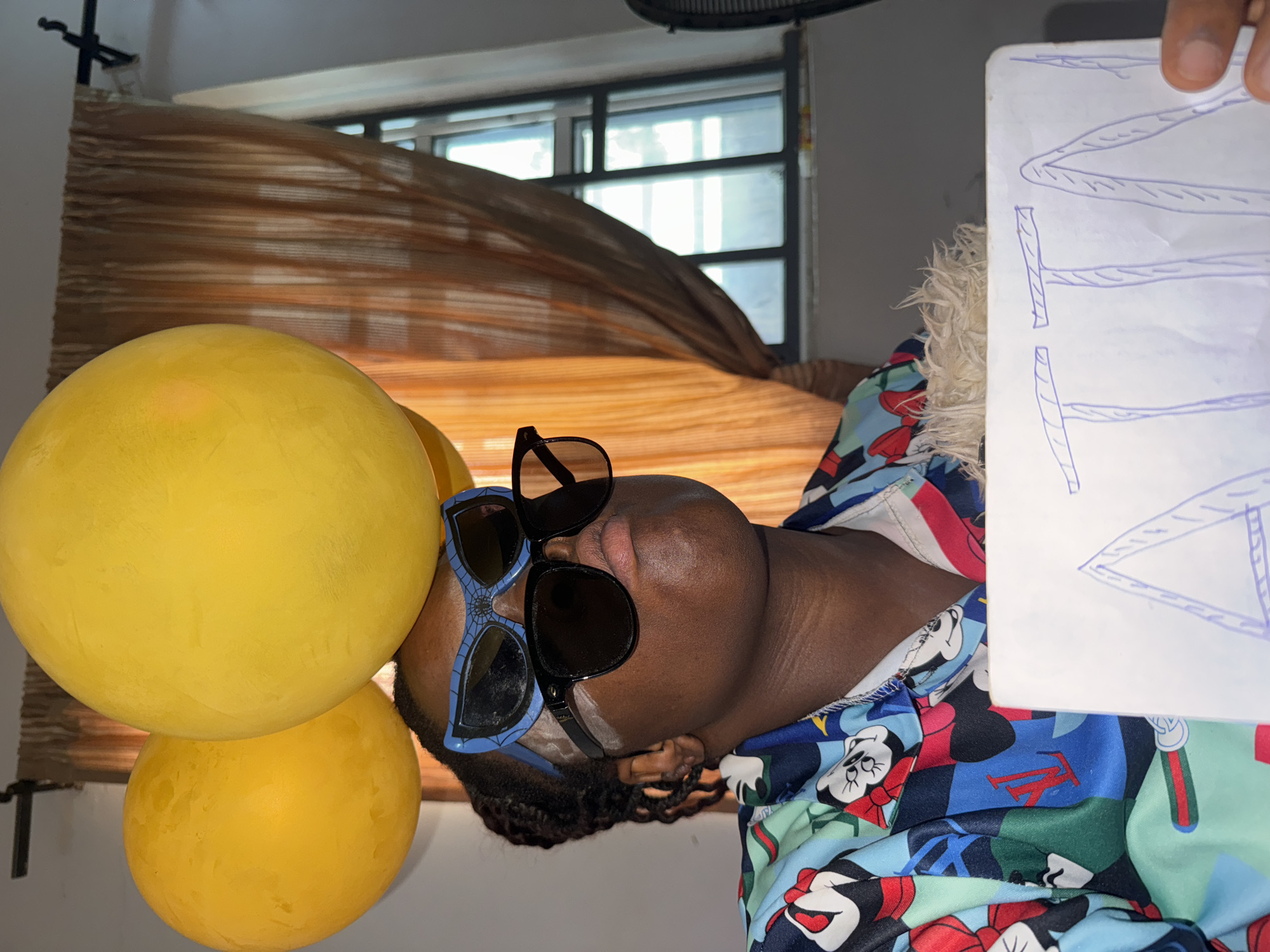 A person lies on their side in front of a brown window shade. They are wearing two pairs of sunglasses, one black and one blue. Two yellow balloons are near the person's head. They are wearing a patterned shirt. A white piece of paper with blue lines is being held in front of them. The location is possibly Sabon Tasha, Nigeria.
